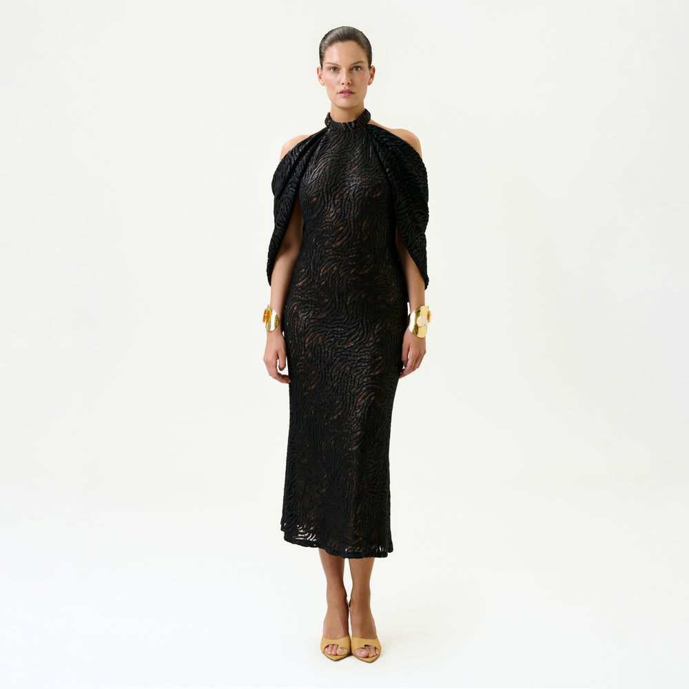 A woman stands facing forward against a plain white background, wearing the Neda Dress—a black, fitted, textured midi dress with cut-out shoulders—paired with gold bracelets and beige open-toe heels. Made in Colombia.