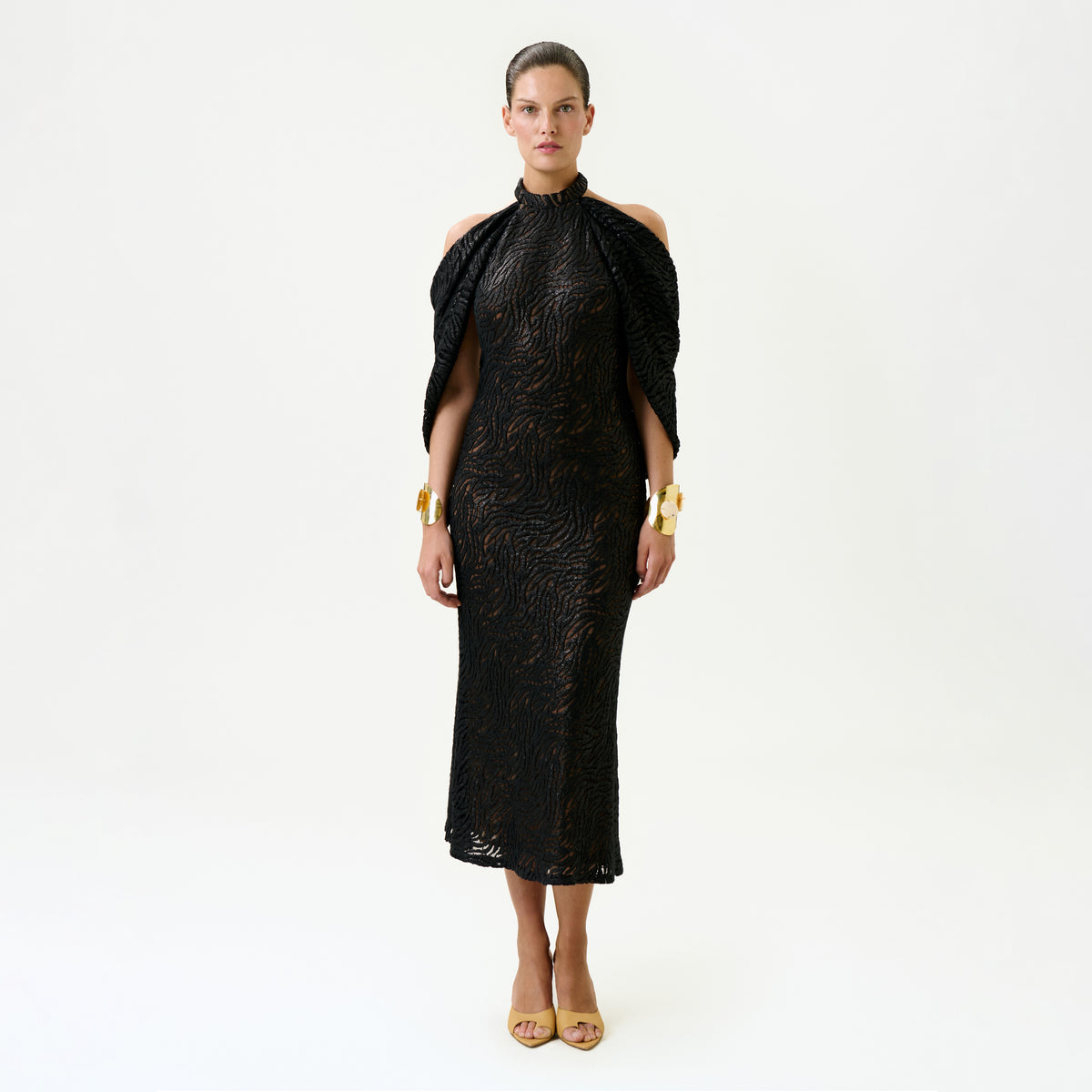 A woman stands facing forward against a plain white background, wearing the Neda Dress—a black, fitted, textured midi dress with cut-out shoulders—paired with gold bracelets and beige open-toe heels. Made in Colombia.