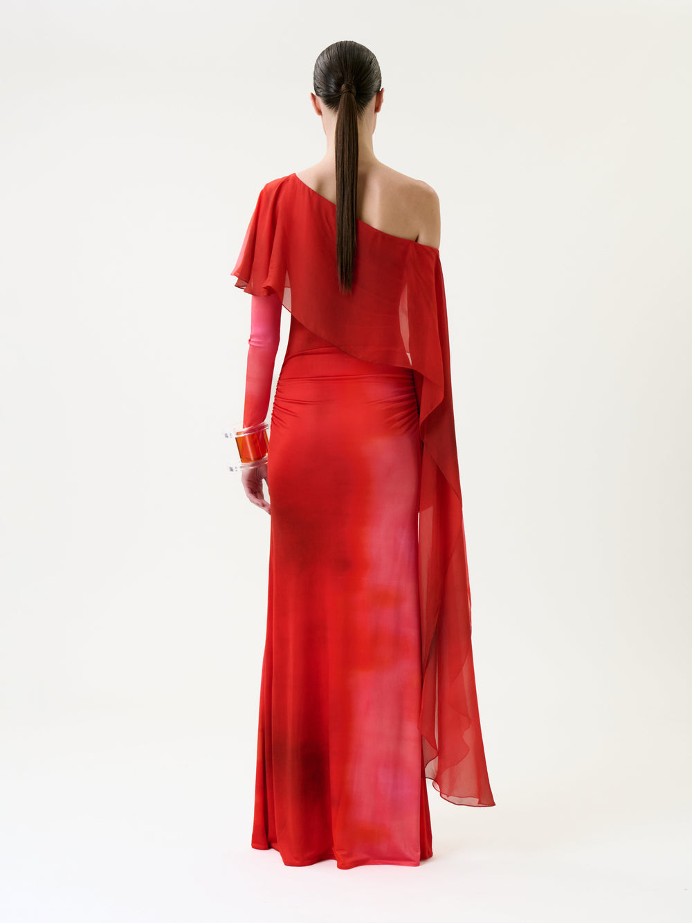 A person with long brown hair in a low ponytail wears the Maddy Dress, a one-shoulder, floor-length red gown featuring sheer draping and translucent red gloves, standing against a plain white background and facing away.