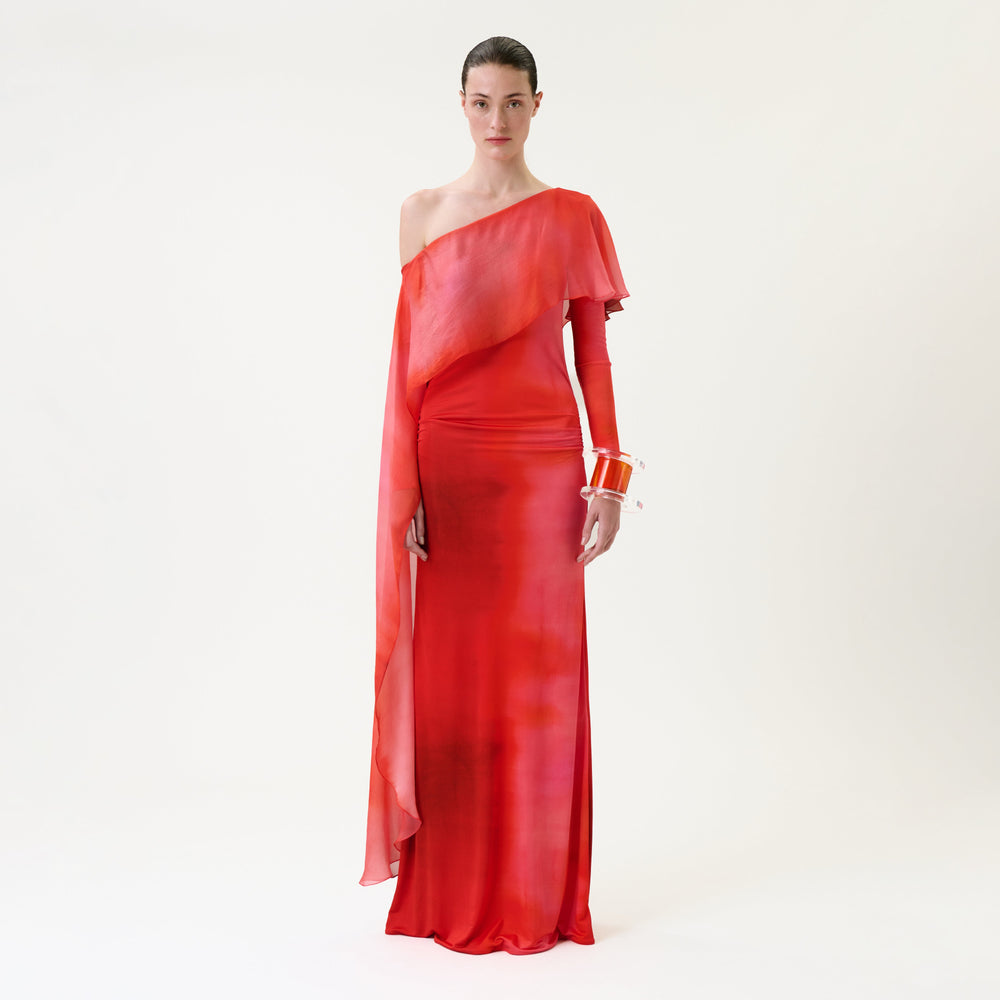 A person stands against a white background wearing the flowing, one-shoulder Maddy Dress—a red gown with an asymmetrical sleeve—accessorized with orange bangle bracelets.