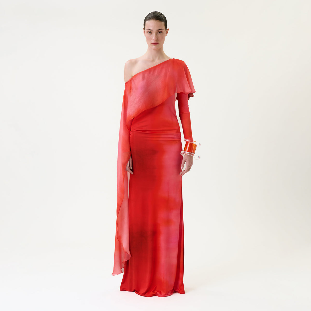 A person stands against a white background wearing the flowing, one-shoulder Maddy Dress—a red gown with an asymmetrical sleeve—accessorized with orange bangle bracelets.