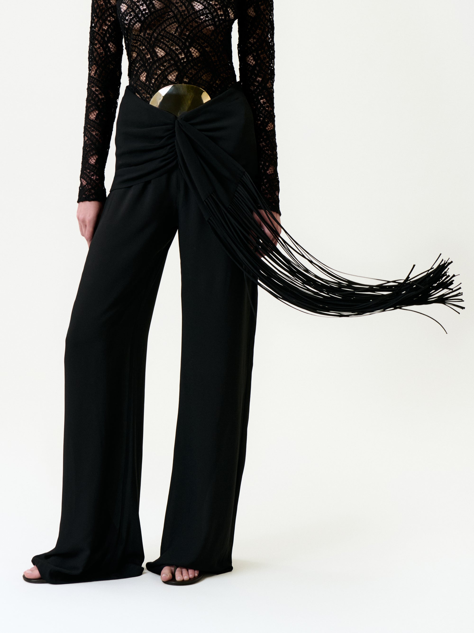 A person stands against a plain white background wearing the Kiele Pant—black wide-leg pants made from 100% Italian Viscose—with a metallic belt, long fringe details, and a black lace long-sleeve top.