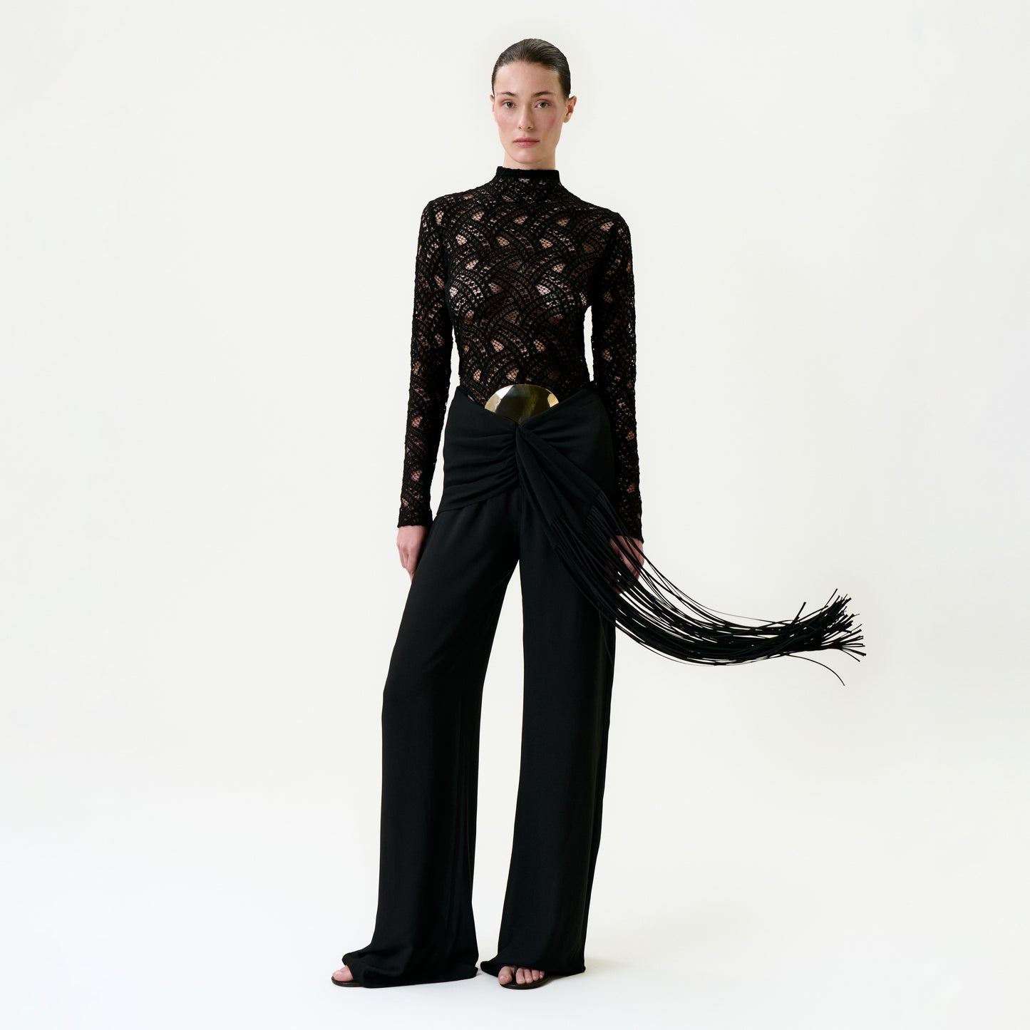 A woman stands against a plain white background wearing the Kiele Pant—high-waisted black pants with a large metallic belt and draped sash of long, flowing fringes, crafted from luxurious Italian fabric.