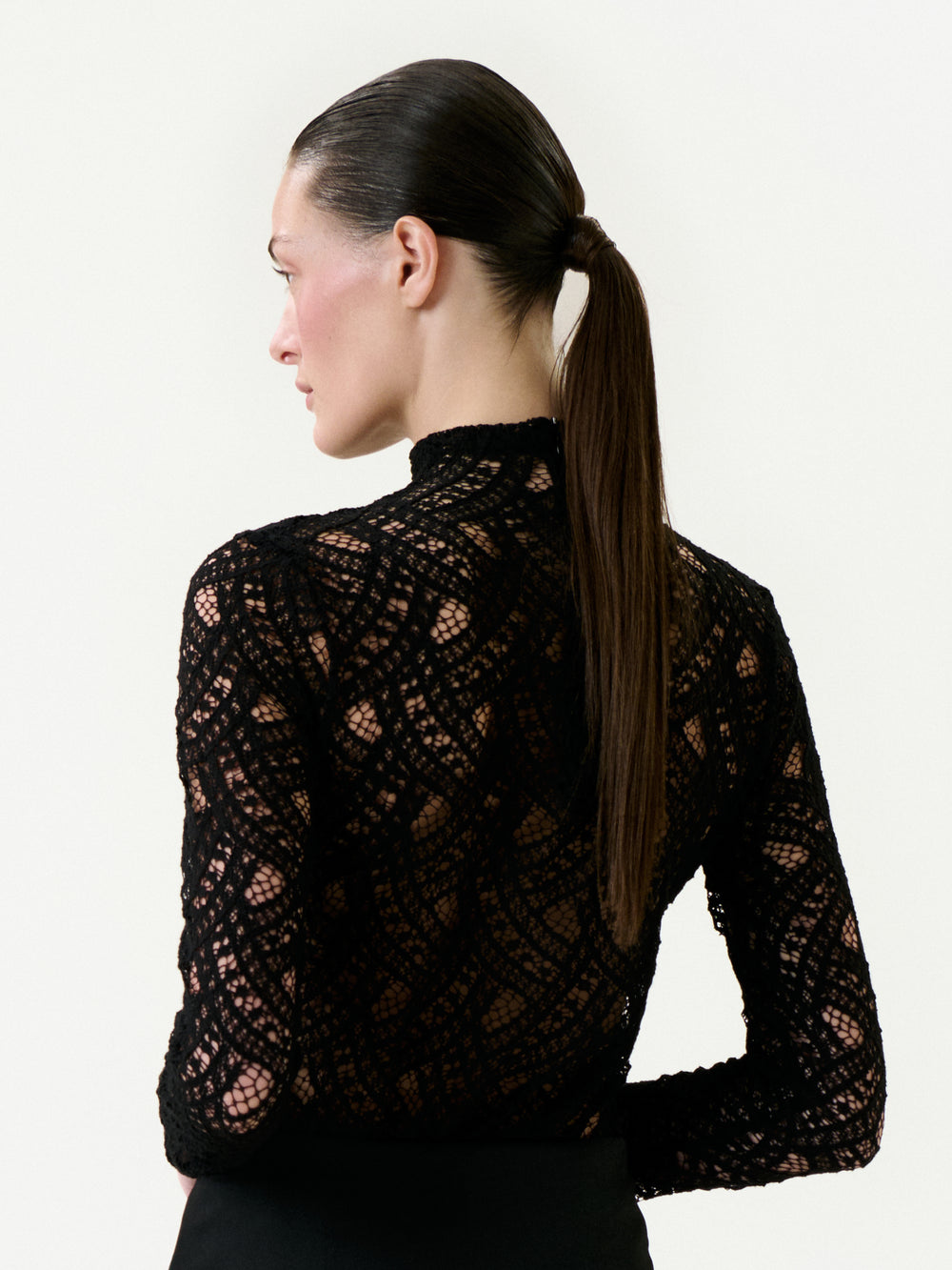 A woman with straight brown hair in a low ponytail is seen from the side and back, wearing the Liz Blouse—a high-neck, long-sleeved black lace top with an intricate Italian pattern. The background is plain white.