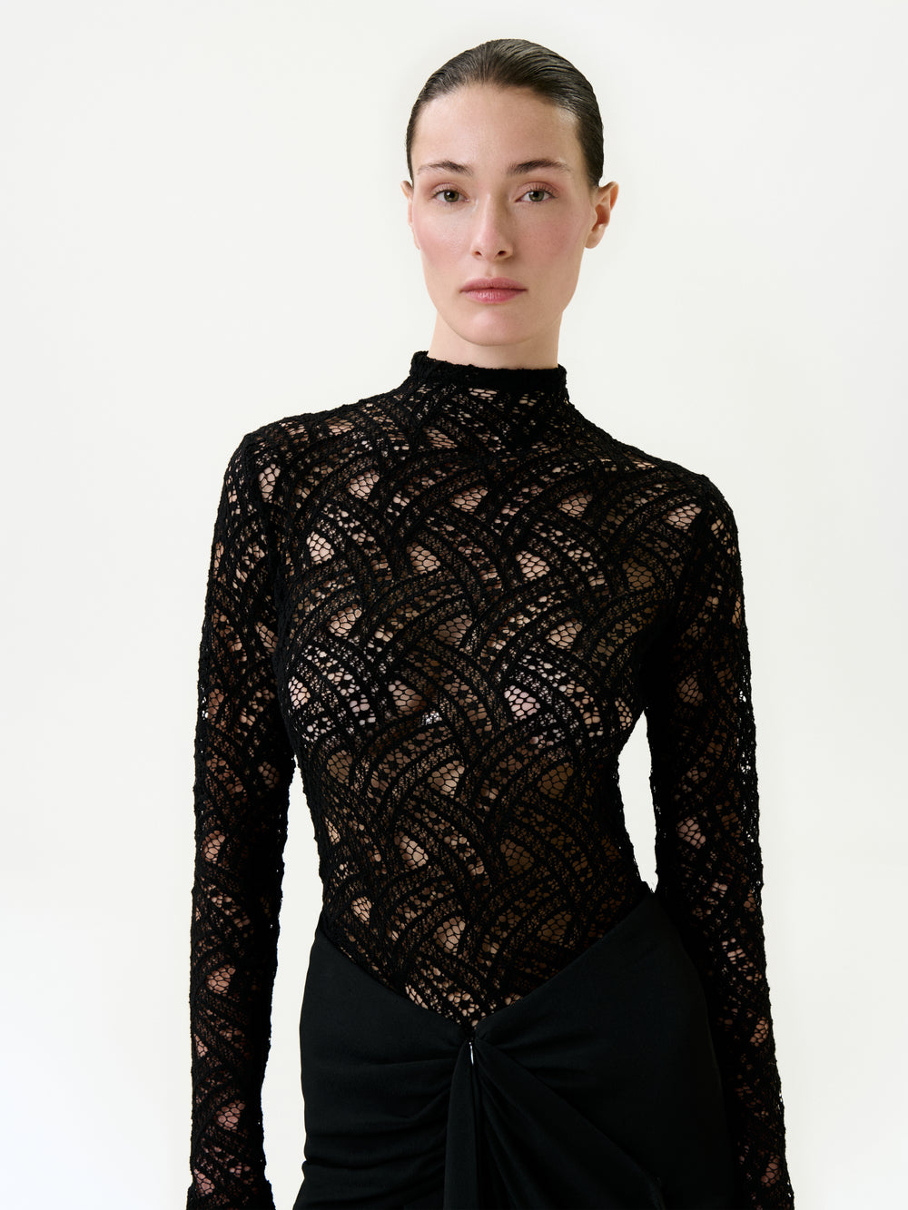 A woman with slicked-back hair wears the Liz Blouse, a black, long-sleeve, cotton-blend lace top featuring a geometric pattern and high neckline. Made in Colombia, she stands against a plain background with a neutral expression.