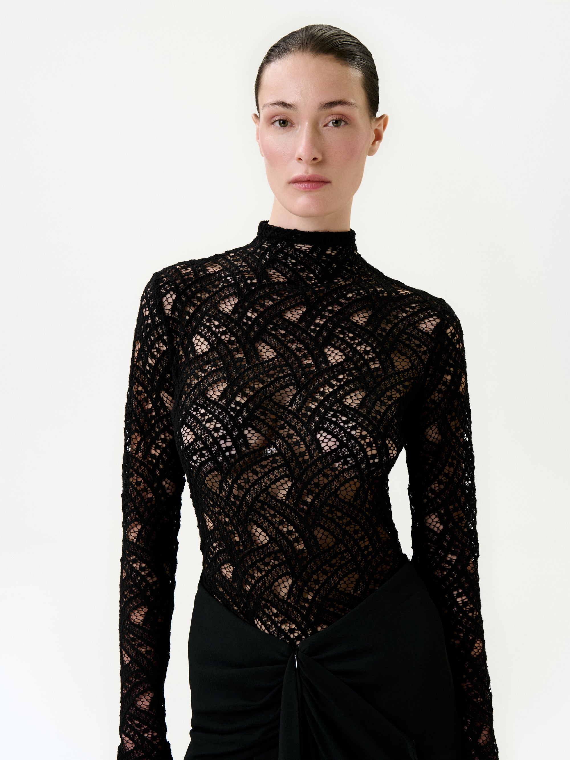 A woman with slicked-back hair wears the Liz Blouse, a black, long-sleeve, cotton-blend lace top featuring a geometric pattern and high neckline. Made in Colombia, she stands against a plain background with a neutral expression.