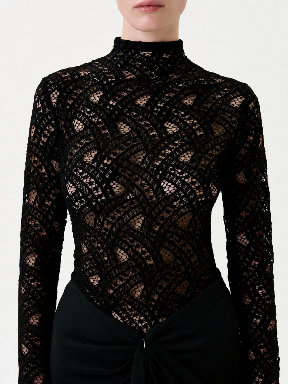 A woman wearing the Liz Blouse—a high-neck, long-sleeve black lace top with a geometric pattern, crafted from premium Italian fabric—paired with a black skirt featuring gathered detail at the waist. Her face is partially out of frame.