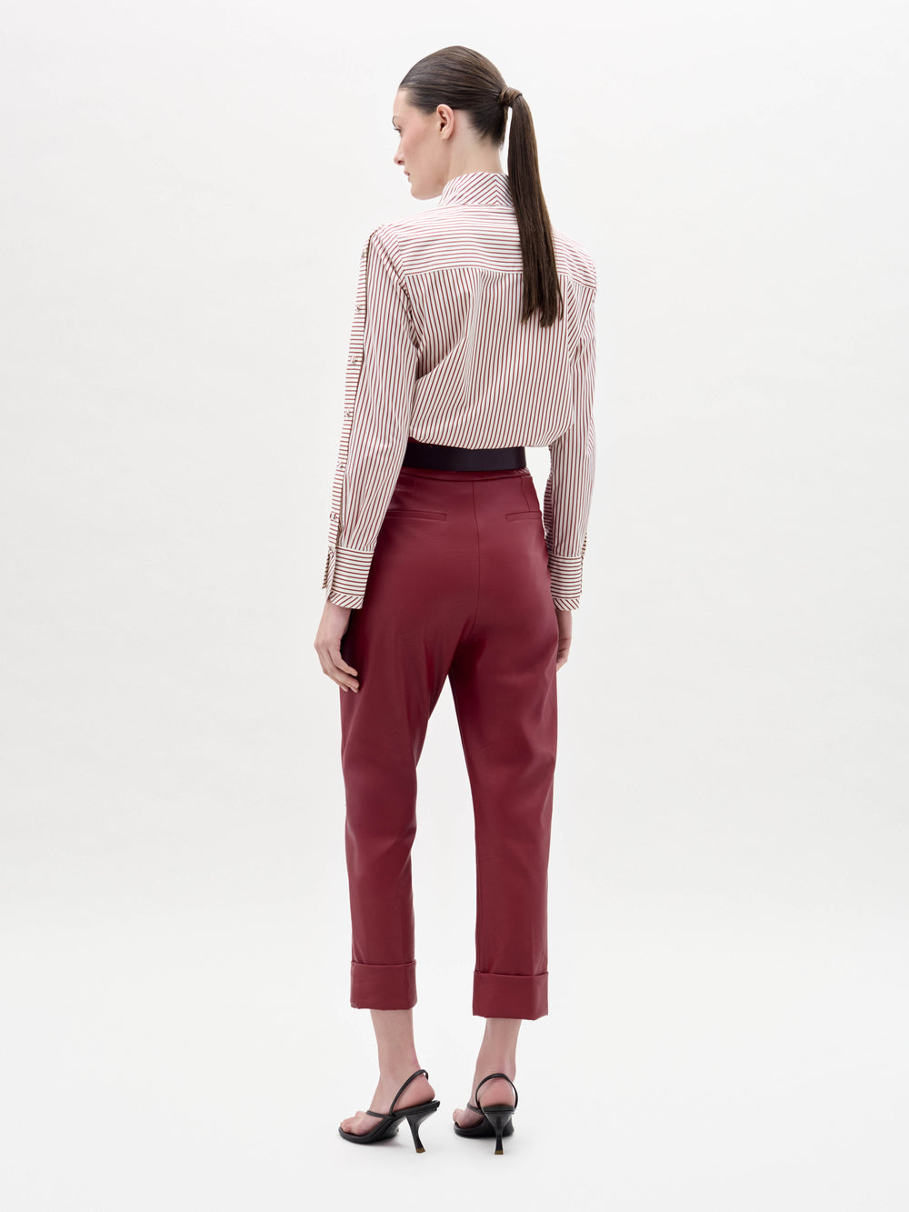 A woman stands against a plain background wearing high-waisted red Moad Pants, a striped shirt with a large red flower applique, a wide belt, and black heeled sandals.