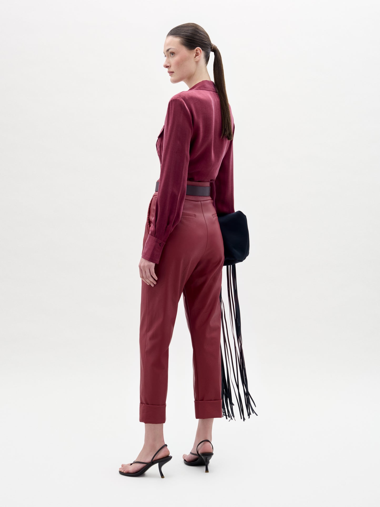 A woman stands sideways wearing the Moad Pant, a pair of high-waisted, cropped pants with a straight fitted leg made from luxurious Italian fabric. She pairs them with a red blouse, black sandals, and carries a black bag adorned with long fringe. Her hair is tied back in a ponytail against a plain white background.