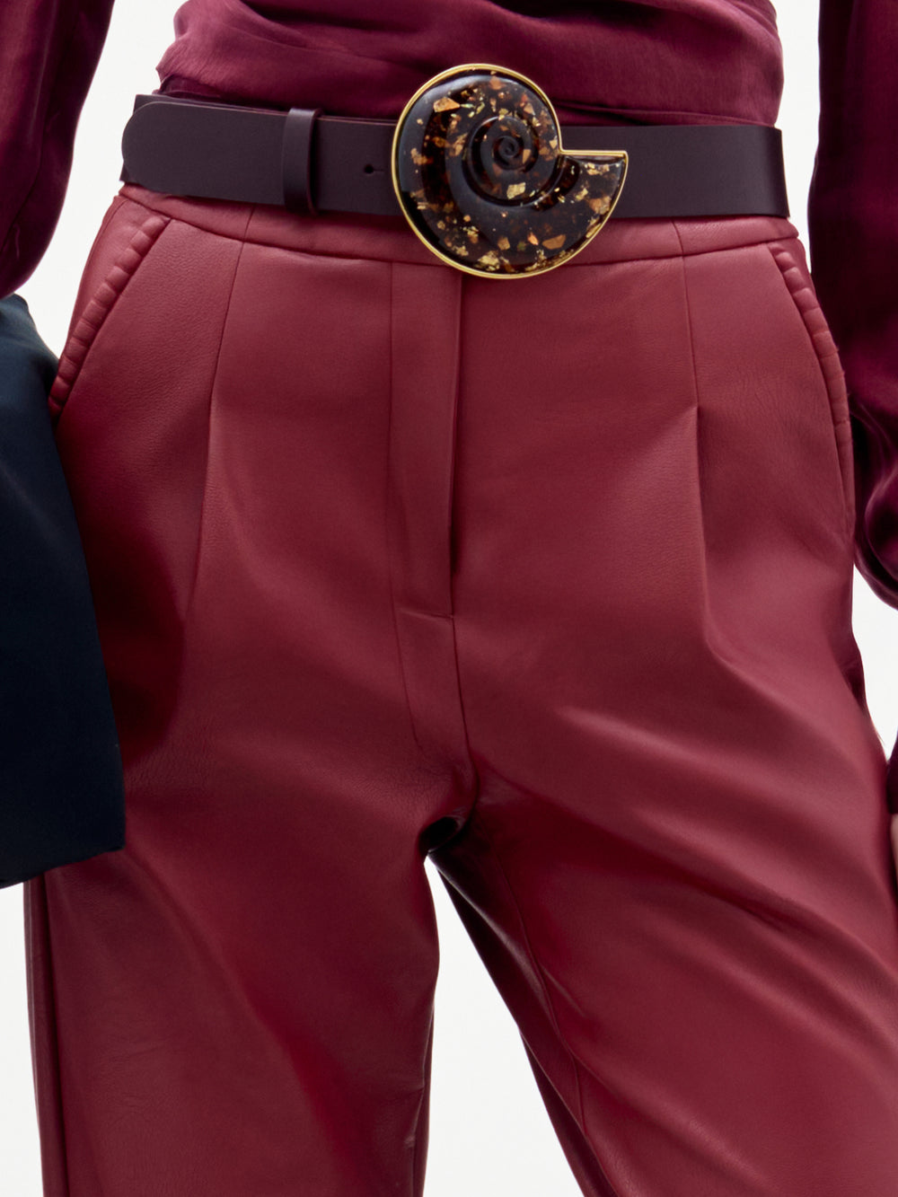 A person wearing the high-waisted, straight-leg Moad Pant in maroon leather with a dark belt featuring a large, spiral-shaped tortoiseshell buckle. The shirt is also maroon, and one hand is holding a dark garment.