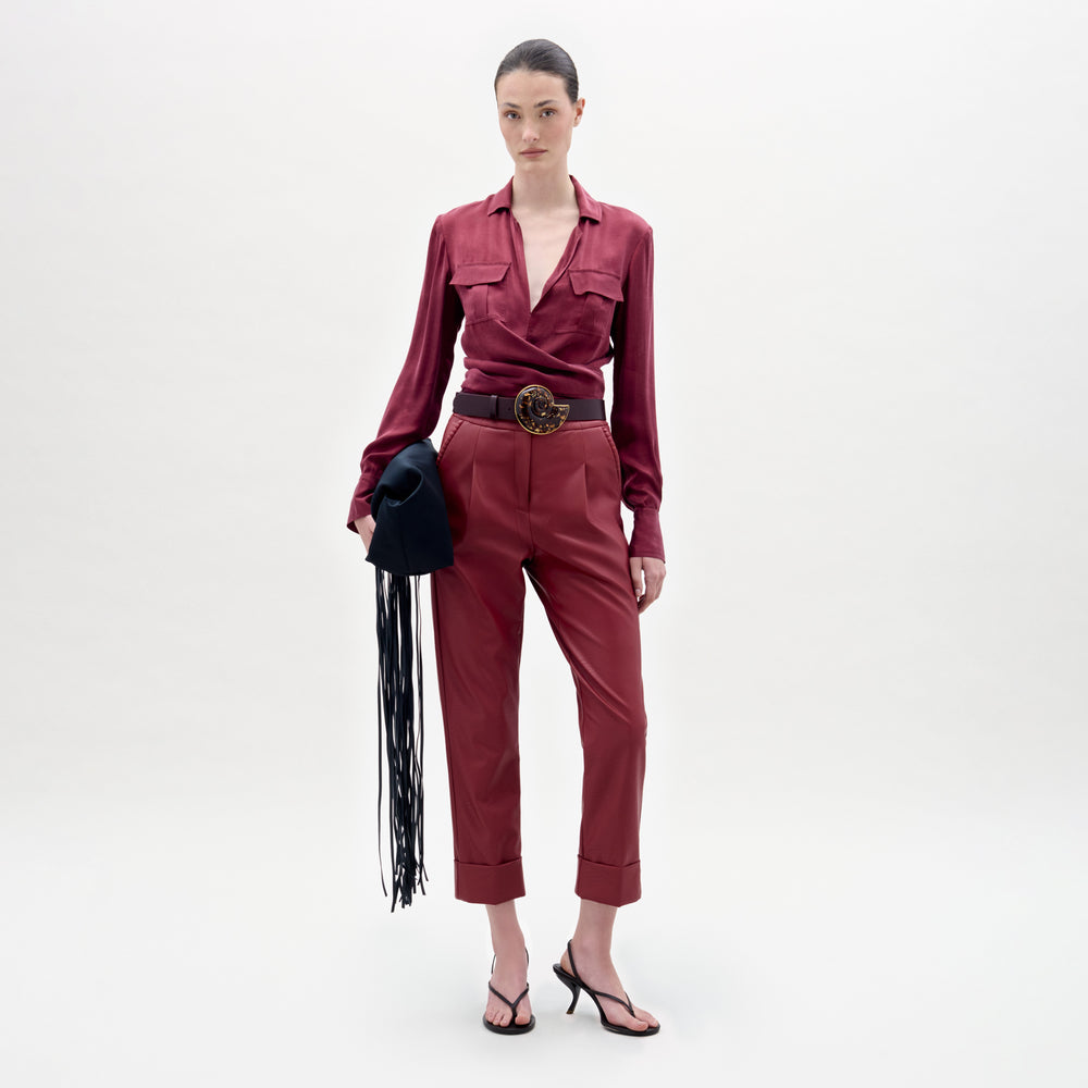 A woman stands against a plain background, wearing a maroon blouse and the Moad Pant—high-waisted, cropped trousers crafted from luxurious Italian fabric. The straight fitted leg is accentuated by a wide belt and heeled sandals, and she completes her look by holding a black hat with fringe.