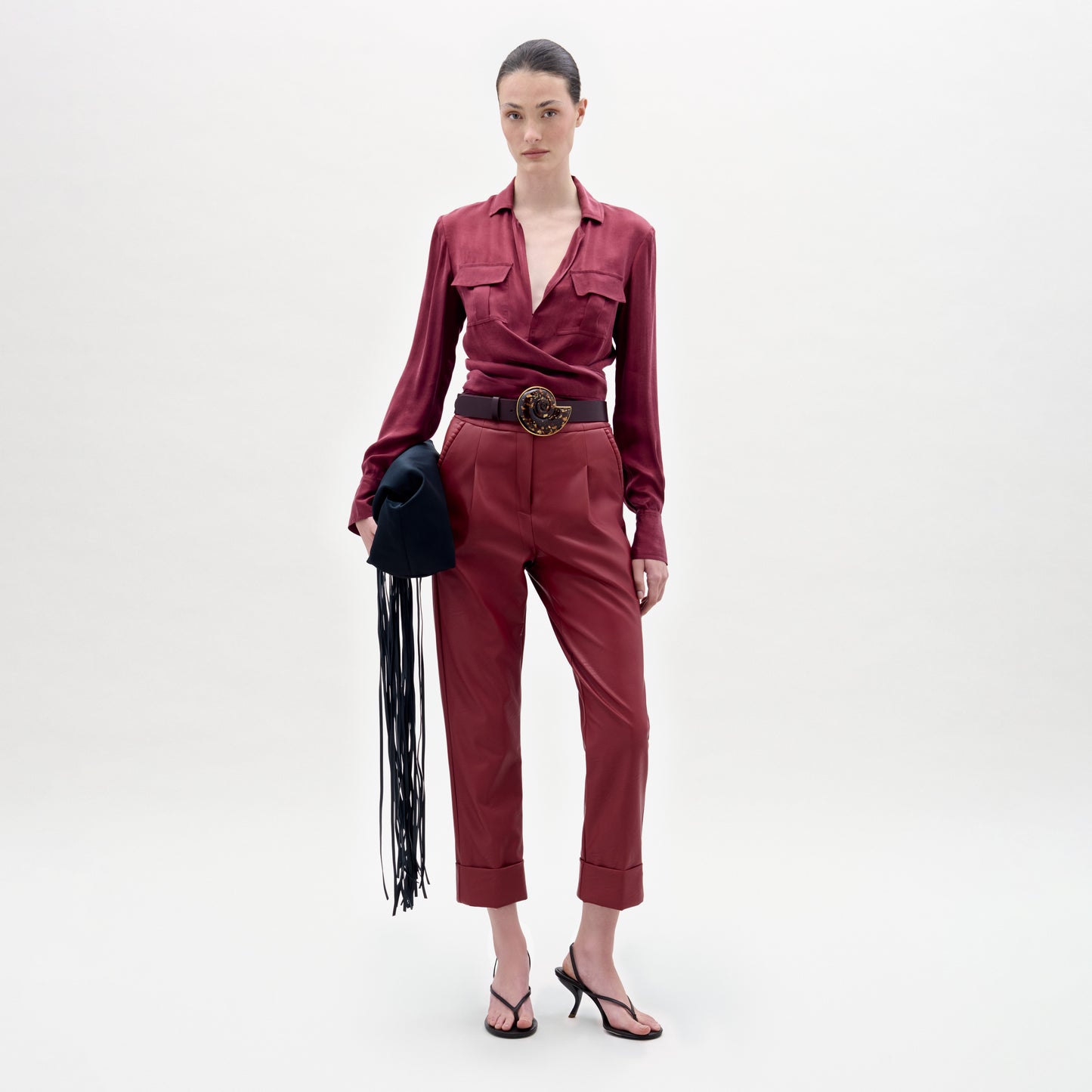 A woman stands against a plain background, wearing a maroon blouse and the Moad Pant—high-waisted, cropped trousers crafted from luxurious Italian fabric. The straight fitted leg is accentuated by a wide belt and heeled sandals, and she completes her look by holding a black hat with fringe.
