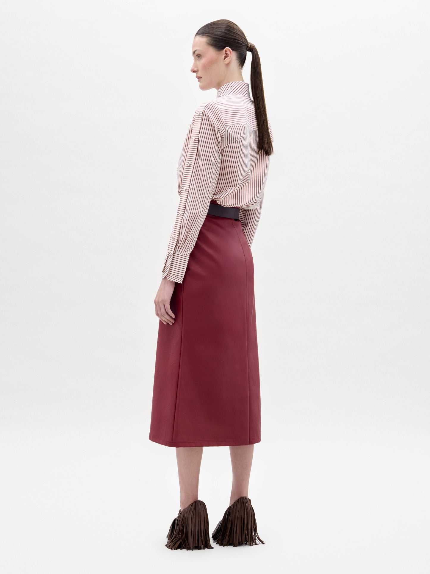 A woman stands against a white background wearing a striped blouse with red floral appliqué, the Layan Skirt in red, a wide belt with a large buckle, and textured brown shoes.