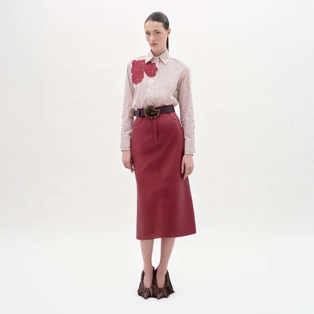 A woman stands against a white background wearing a striped blouse with red floral appliqué, the Layan Skirt in red, a wide belt with a large buckle, and textured brown shoes.