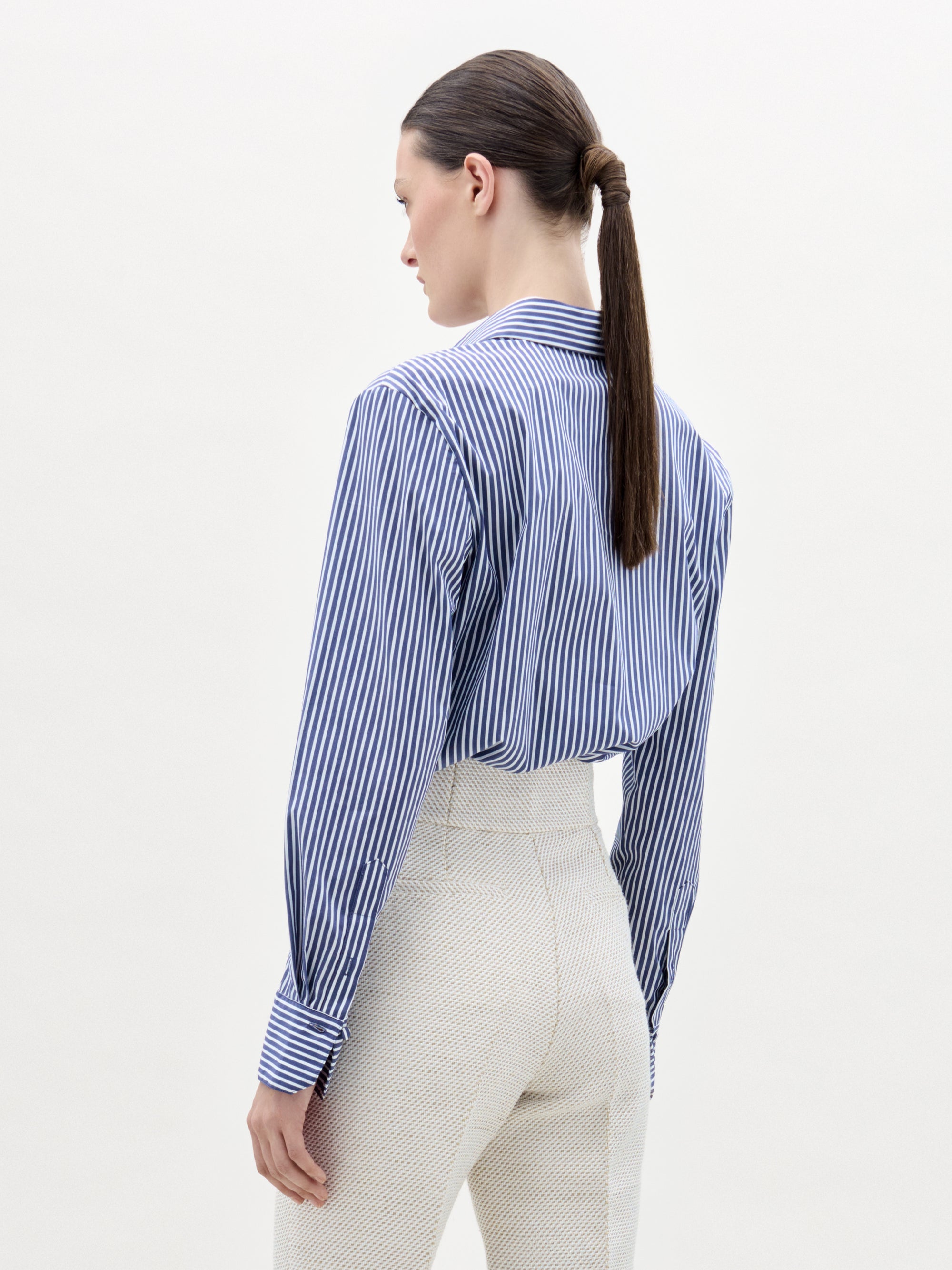 A woman with a ponytail wears the Karole Blouse, a striped button-down shirt crafted from Italian fabric.