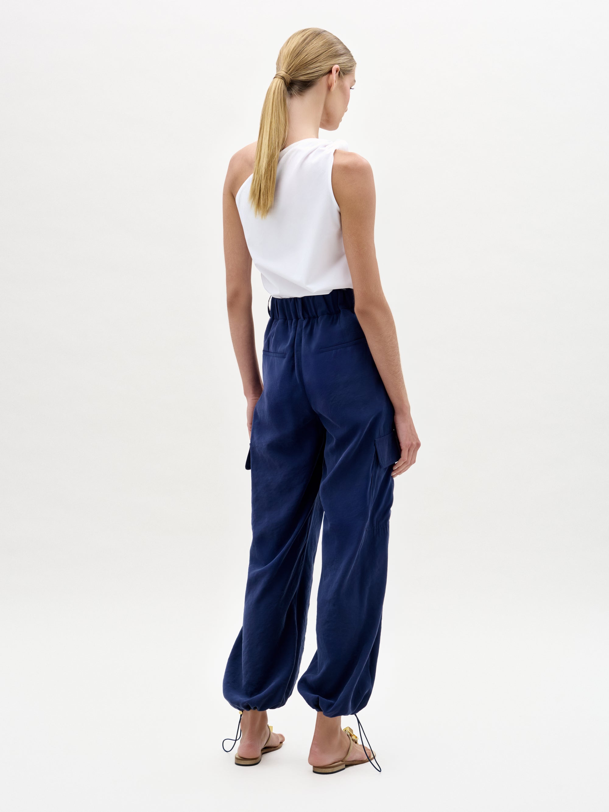 A woman with blonde hair in a low ponytail wears a sleeveless white top and the Darnell Pant—loose, high-waist navy blue cargo pants with a drawstring hem, made from fine Italian fabric. She stands facing away against a plain light background in flat sandals.
