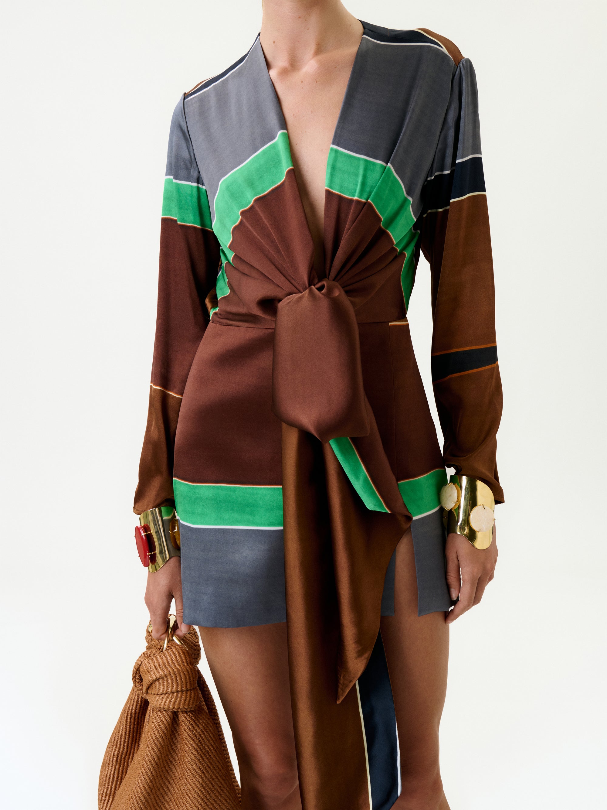 A person wears the Midori Dress—an Italian-fabric, long-sleeve mini dress with green, brown, and gray color blocks plus a large front knot. They carry a textured brown bag and don chunky gold bracelets against a white background.