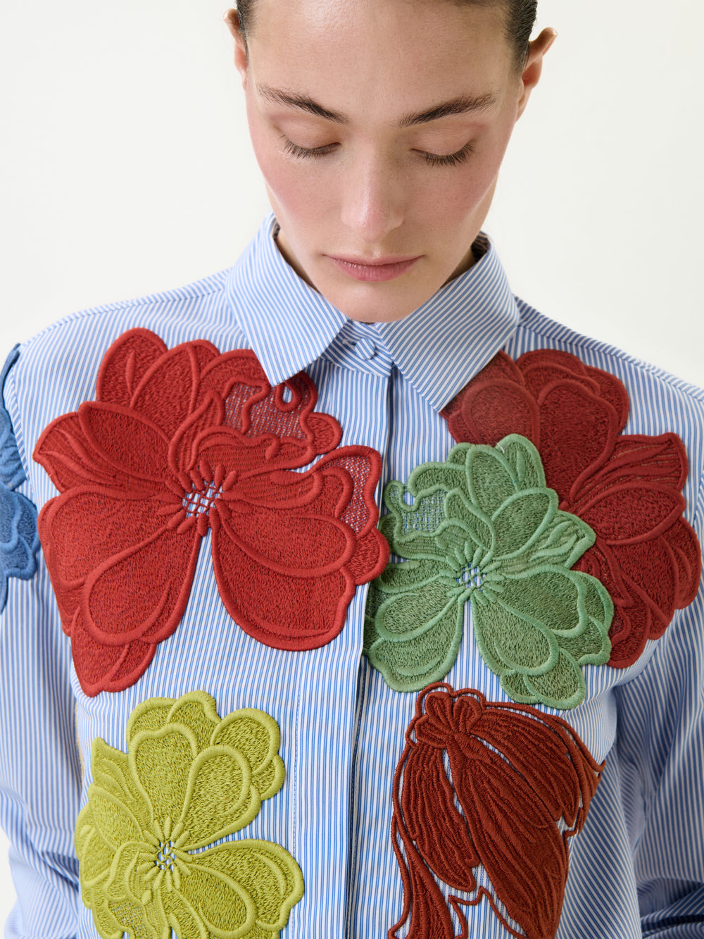 Wearing the Warda Shirt, a blue and white striped 100% cotton top featuring large red, green, and yellow floral appliqués, the person looks down with a neutral expression.