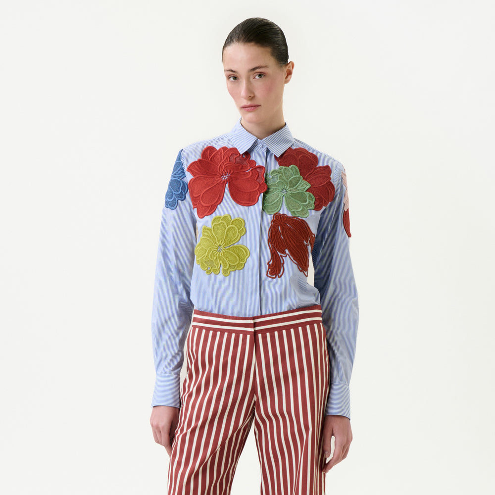 A person wears the Warda Shirt, a light blue 100% cotton top with large, colorful floral appliqués, paired with red-and-white striped pants, standing against a plain white background.