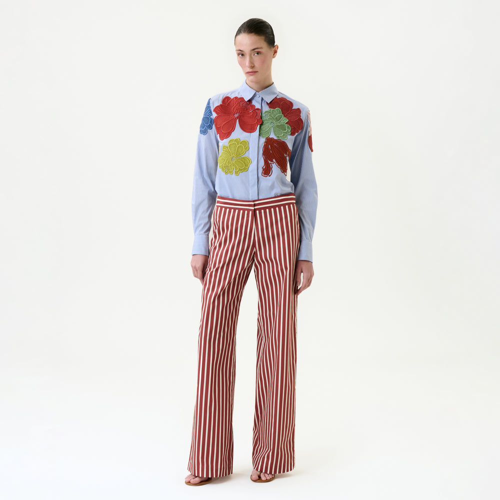 She wears the Ulla Pant, featuring high-waisted red-and-white vertical stripes and a wide-leg silhouette, paired with a light blue shirt adorned with colorful leaf appliqués. Her hair is pulled back and she completes the look with sandals.