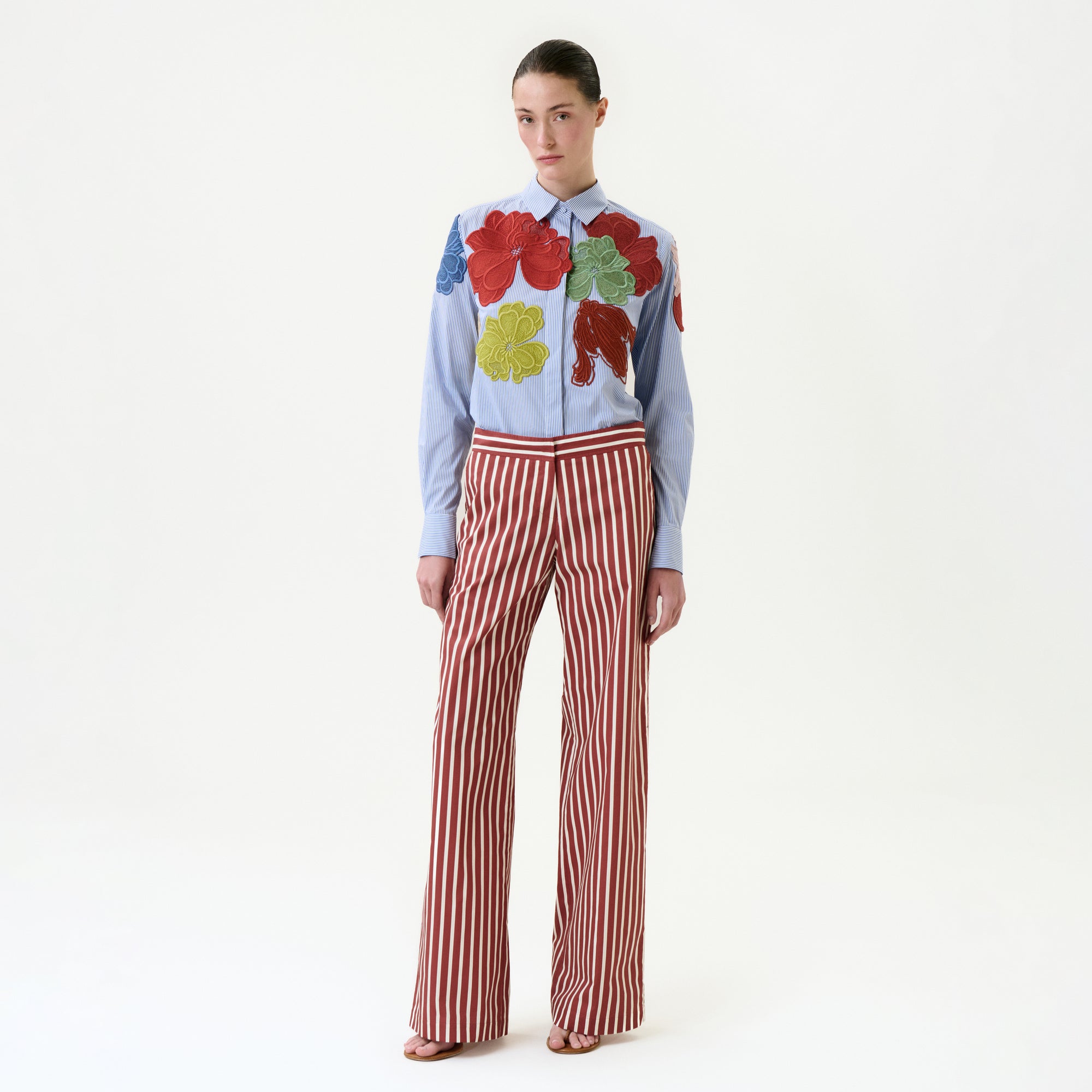 She wears the Ulla Pant, featuring high-waisted red-and-white vertical stripes and a wide-leg silhouette, paired with a light blue shirt adorned with colorful leaf appliqués. Her hair is pulled back and she completes the look with sandals.