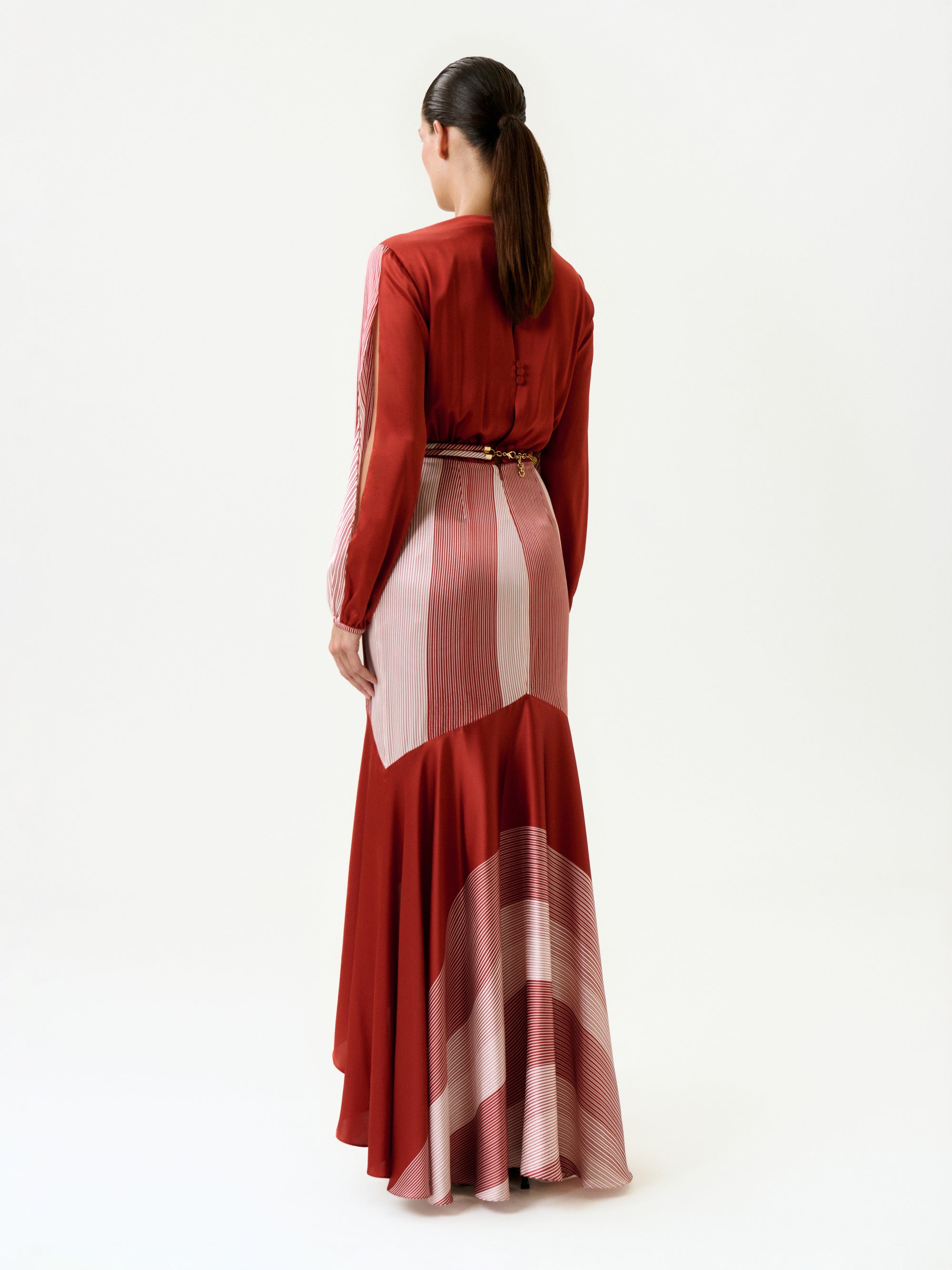 A woman with a ponytail stands with her back to the camera, wearing the Mairet Dress—a long, flowing silk maxi dress with geometric patterns and a belted waist—set against a plain white background.