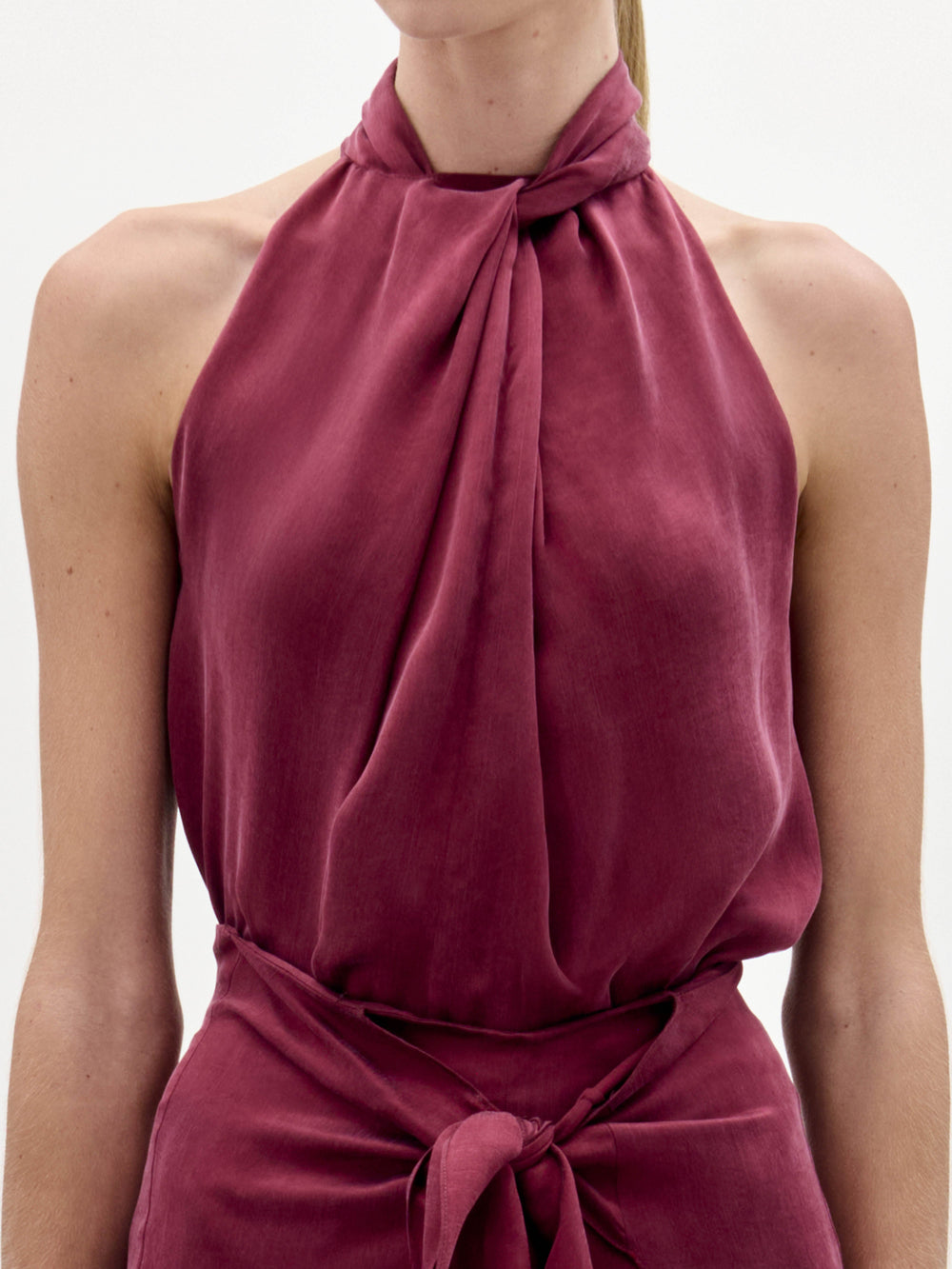 A woman stands against a plain background wearing the Shela Blouse, a sleeveless, high-neck magenta top with a tie detail at the waist, paired with a small handbag.