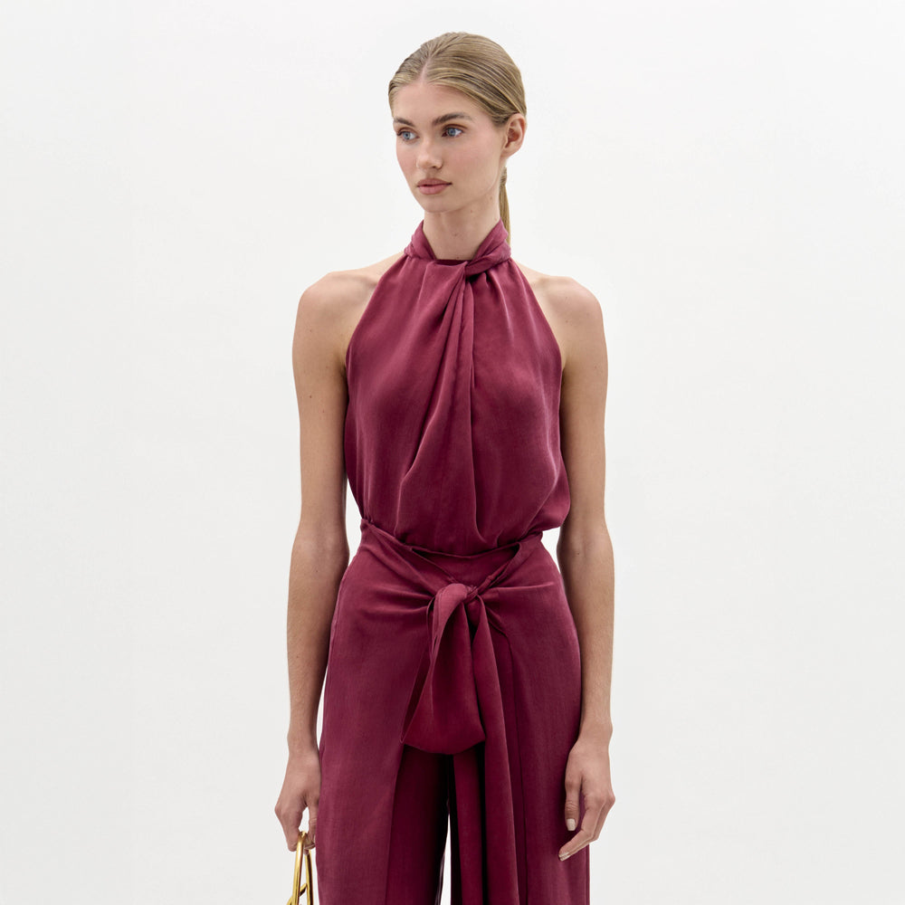 A woman stands against a plain background wearing the Shela Blouse, a sleeveless, high-neck magenta top with a tie detail at the waist, paired with a small handbag.