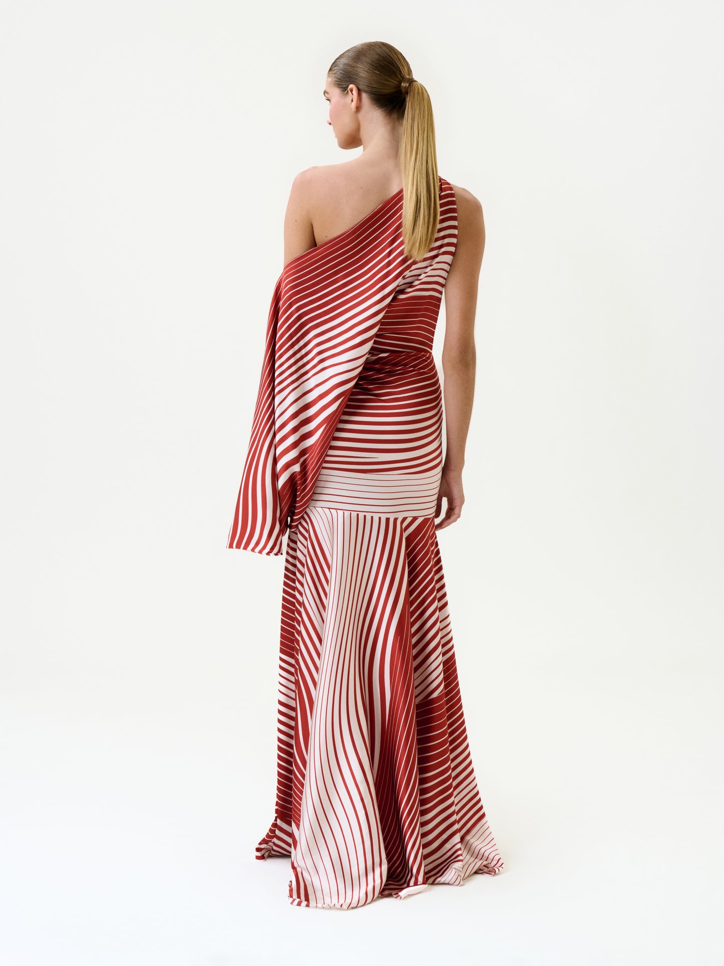 A woman stands facing away in the Noy Dress—a red and white striped silk maxi with a fitted bodice and flowing skirt, crafted from luxurious Italian fabric—her hair in a low ponytail against a plain white background.