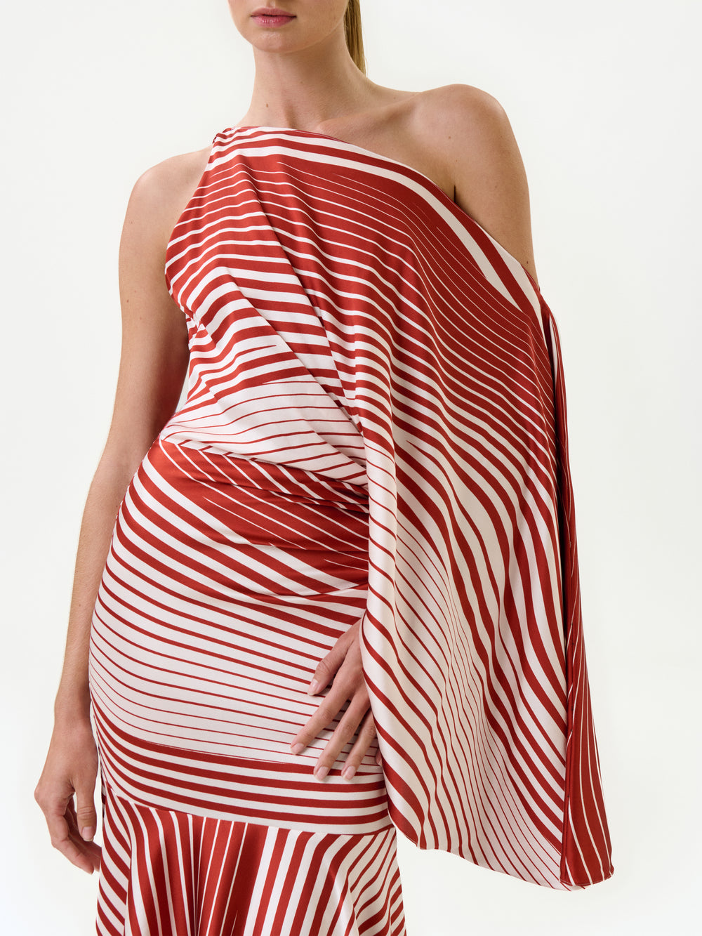 A person wearing the Noy Dress—a red and white striped, one-shoulder maxi dress made from Italian fabric with diagonal and horizontal lines—stands against a white background. Only the upper body and part of the skirt are visible.