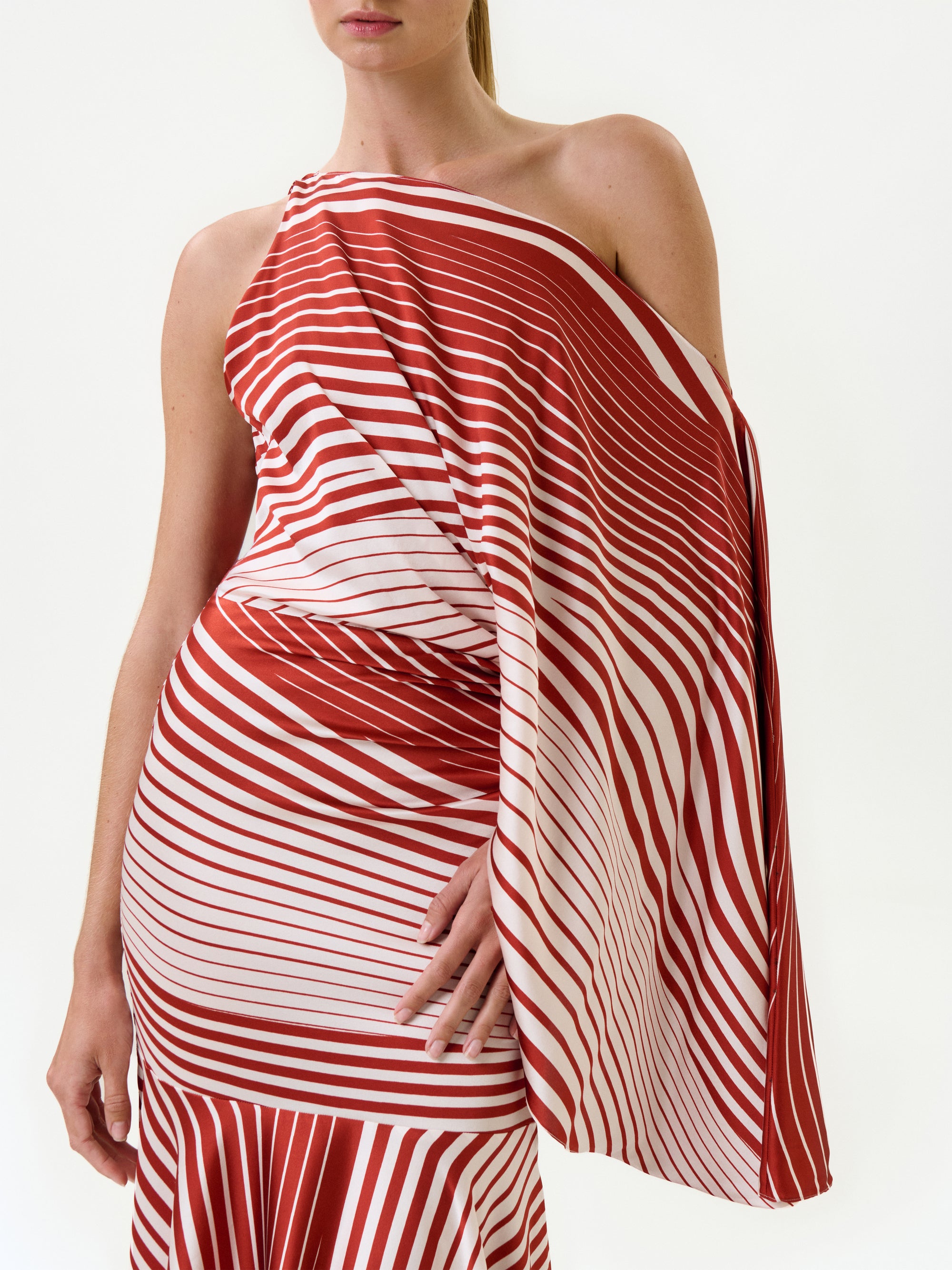 A person wearing the Noy Dress—a red and white striped, one-shoulder maxi dress made from Italian fabric with diagonal and horizontal lines—stands against a white background. Only the upper body and part of the skirt are visible.