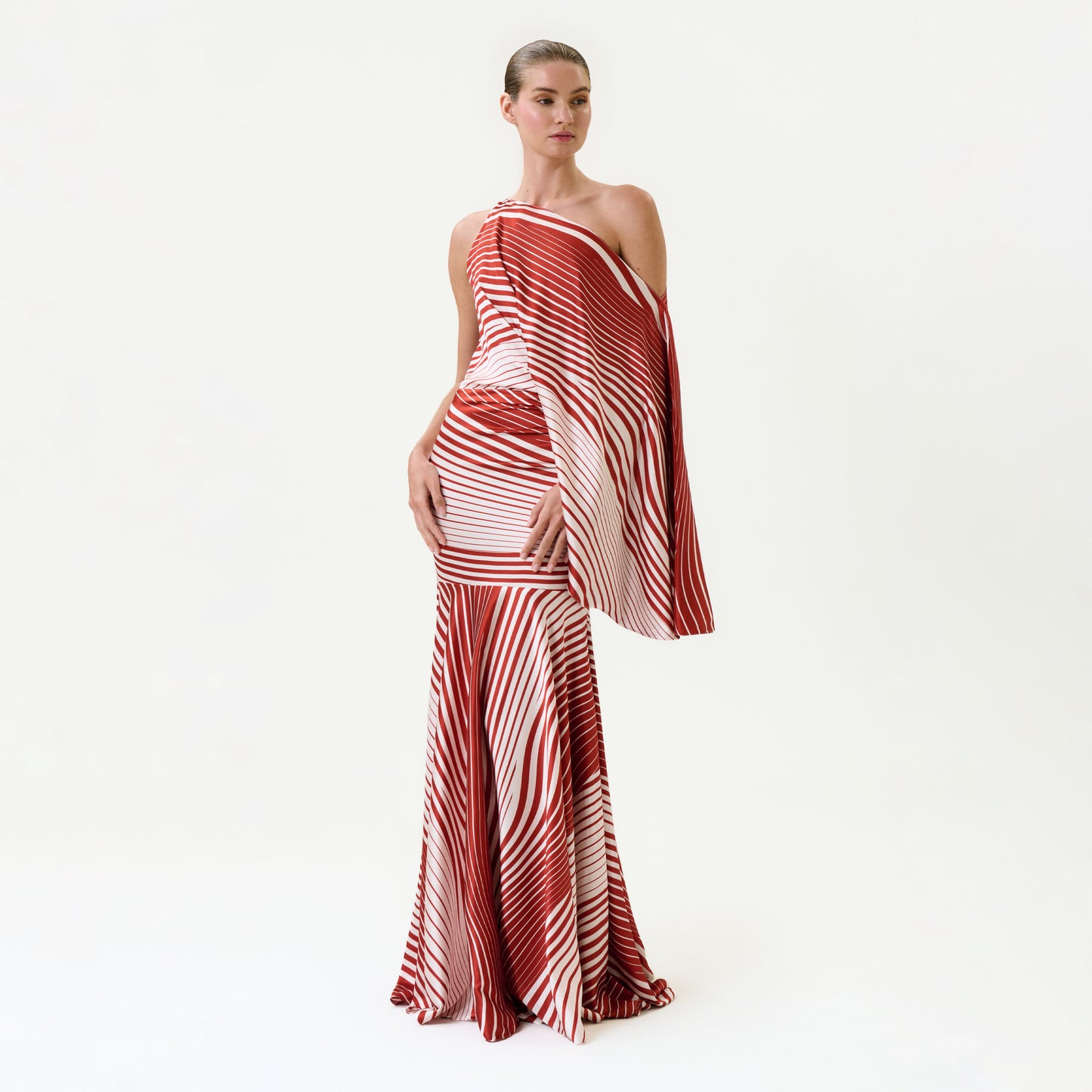 Against a plain background, a person in the Noy Dress—a red and white striped, one-shoulder silk maxi with a draped sleeve and flowing skirt—poses confidently with slicked-back hair.