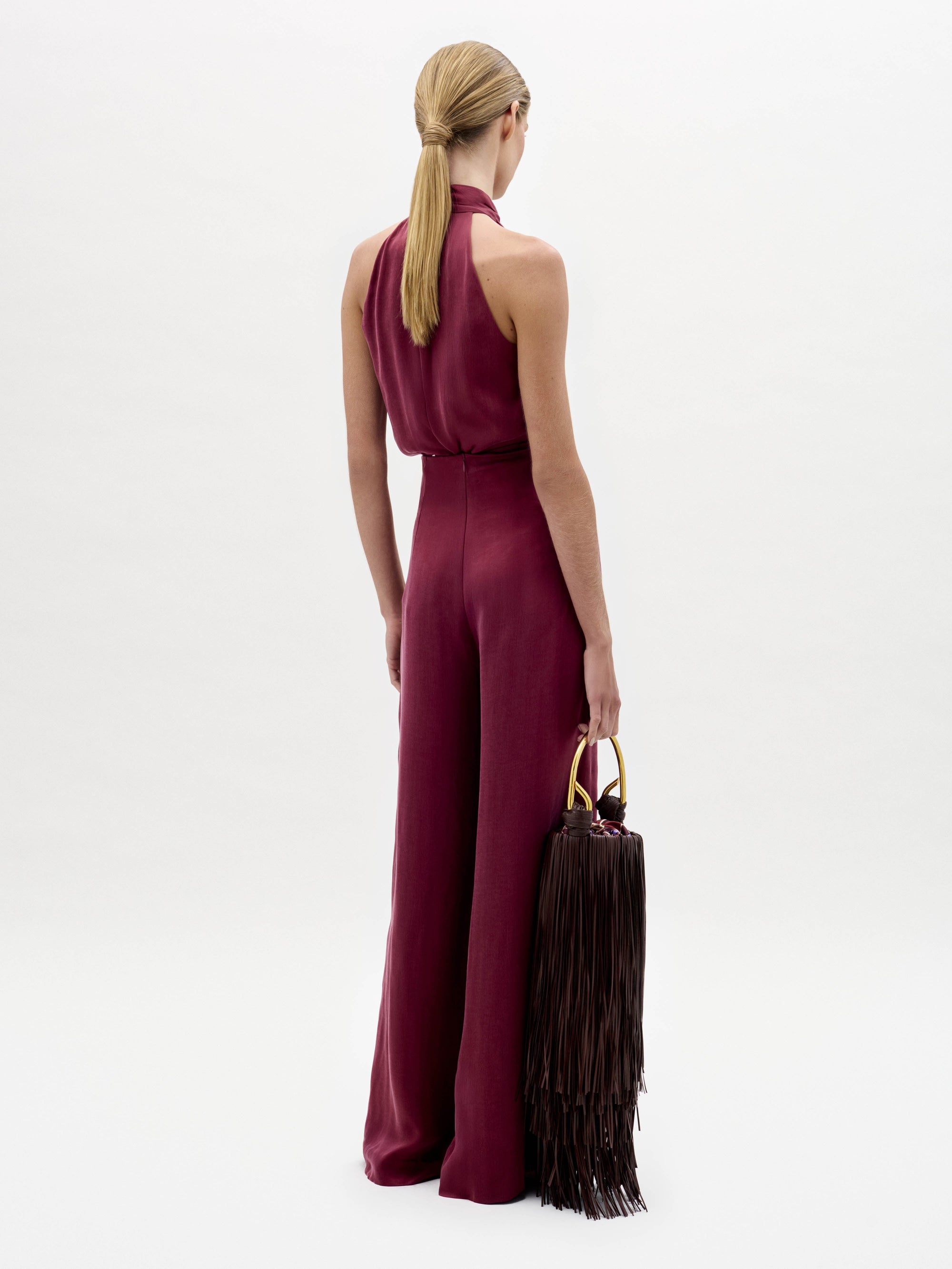 A woman stands against a white background wearing the Nuria Pant—a sleeveless maroon halter-neck jumpsuit with a wide waist tie—while holding a dark fringed handbag.