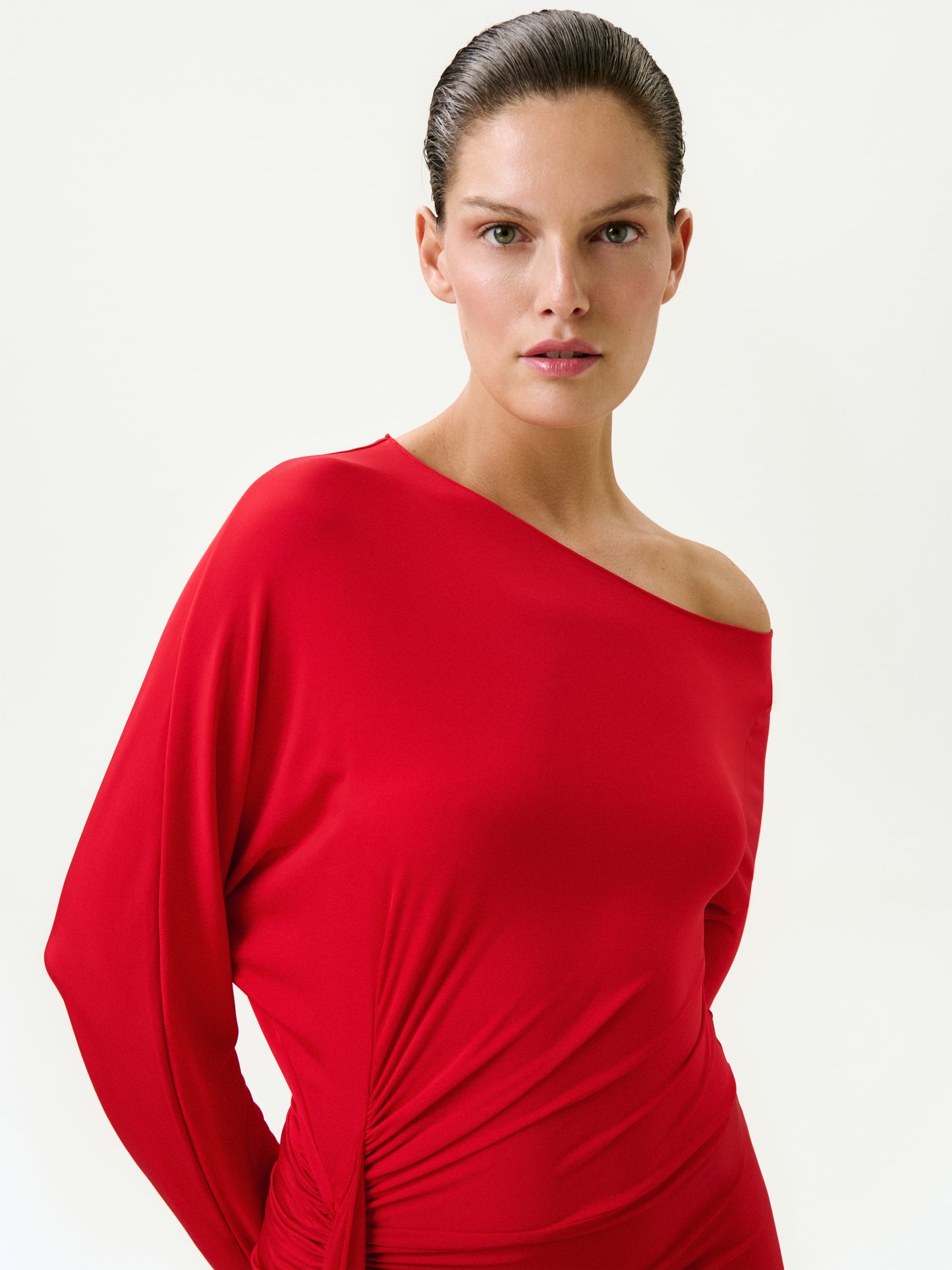 A woman with neat, slicked-back hair wears the Roxy Dress, a vibrant red one-shoulder maxi made in Colombia. She stands against a plain light background, looking directly at the camera with a neutral expression.