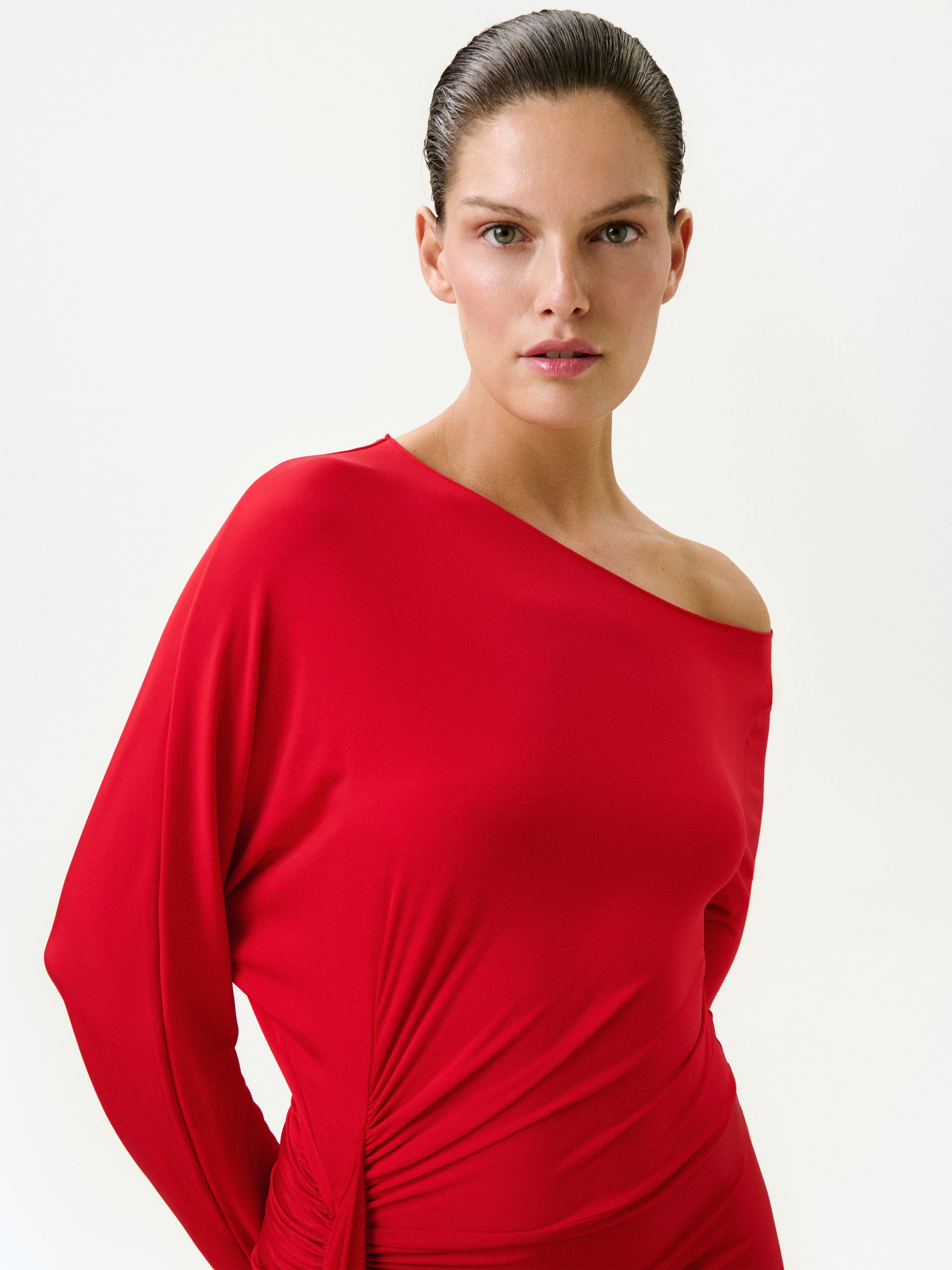 A woman with neat, slicked-back hair wears the Roxy Dress, a vibrant red one-shoulder maxi made in Colombia. She stands against a plain light background, looking directly at the camera with a neutral expression.