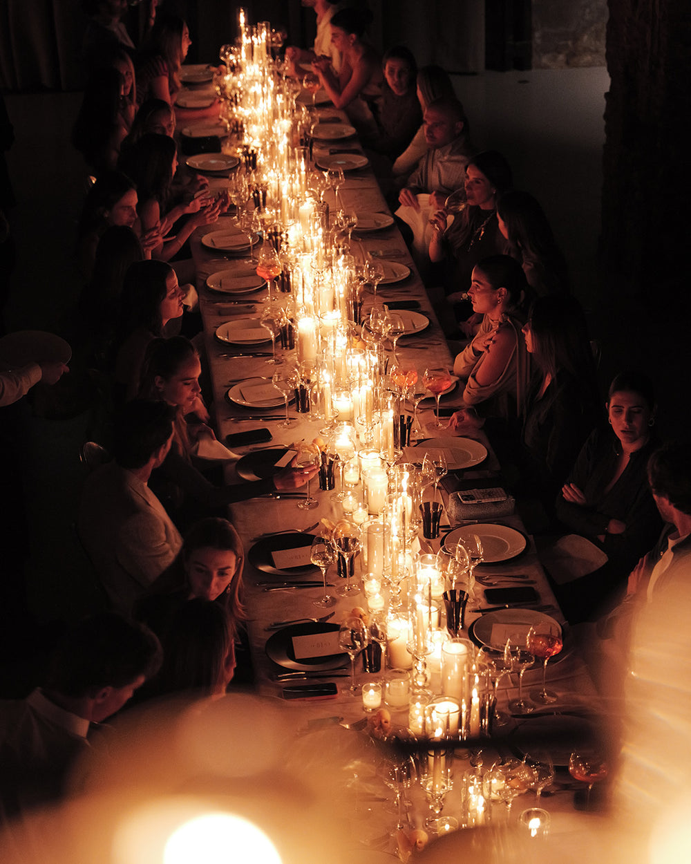 A long dining table set for guests under a dense arrangement of lit candles, creating a warm, atmospheric evening scene in a dimly lit room.
