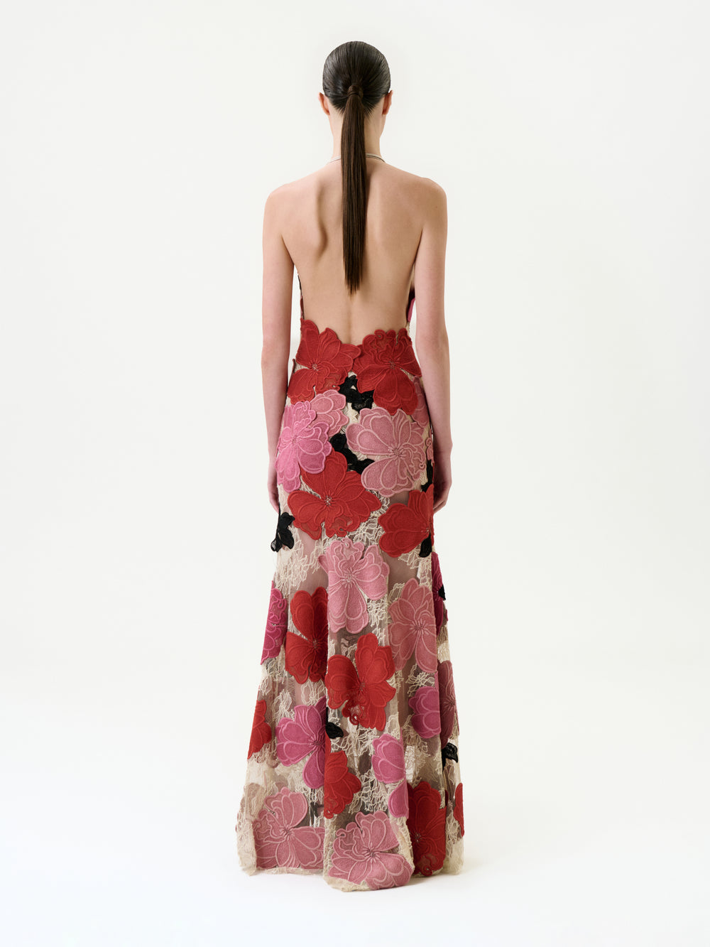 A person with long, dark hair in a low ponytail models the Vanessa Dress—a strapless, backless maxi featuring bold red, pink, black, and beige floral prints on fine Italian fabric—standing against a plain white background.