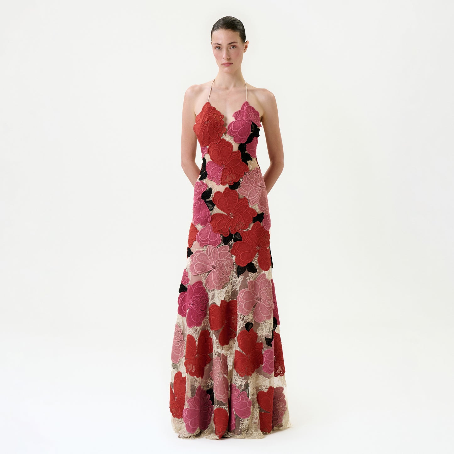 A woman with a neutral expression stands against a white background wearing the Vanessa Dress, a Colombian-made maxi dress with thin straps and bold red, pink, and black floral patterns on a beige base. Her hands are behind her back.