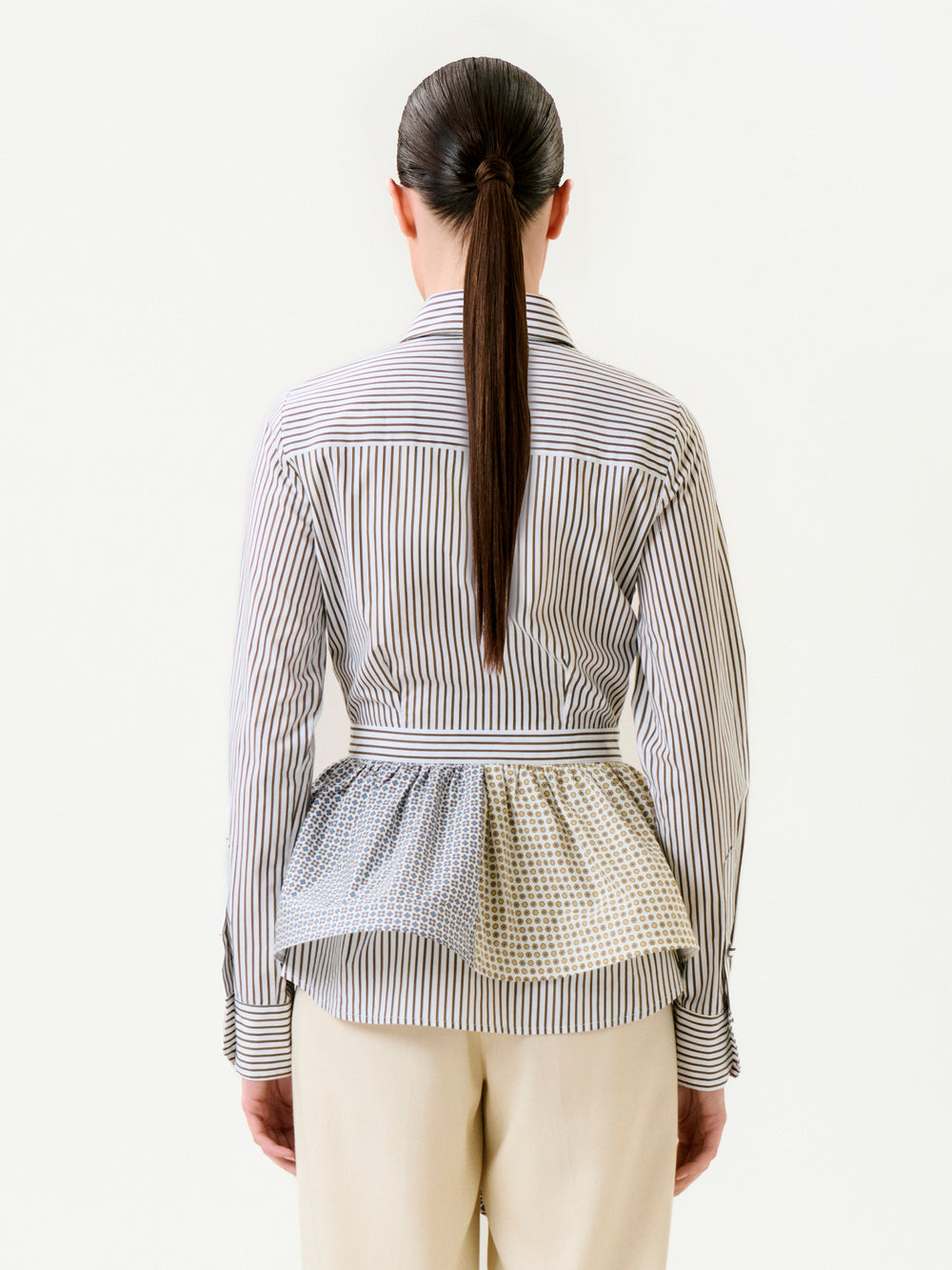 A person with long, straight brown hair in a low ponytail is seen from behind wearing the Malina Shirt—a 100% cotton button-up with peplum detail, crafted from Portuguese fabric with mixed stripe patterns—paired with beige pants.