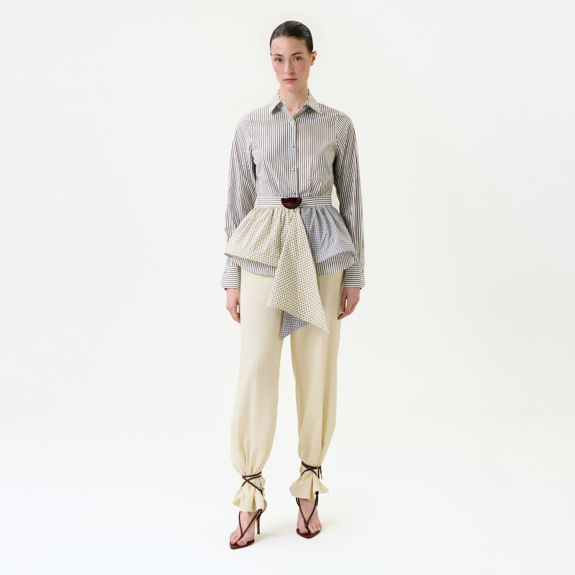A woman with slicked-back hair wears beige tied-cuff pants, heeled sandals, a belted asymmetric peplum overlay, and the Malina Shirt—an elegant striped button-up made from premium Portuguese fabric—against a plain white background.