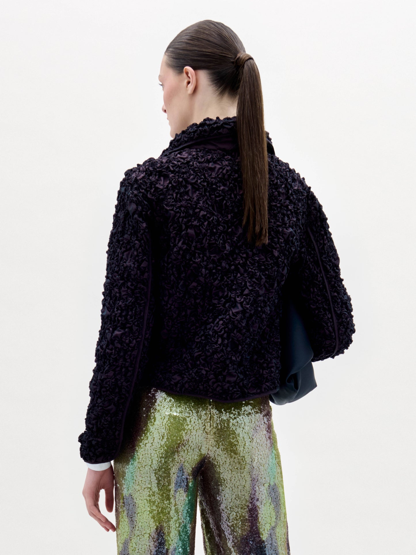 A woman stands against a plain background, wearing the textured black Jamari Jacket over a white shirt and shiny patterned pants, holding a folded black item in her left arm.