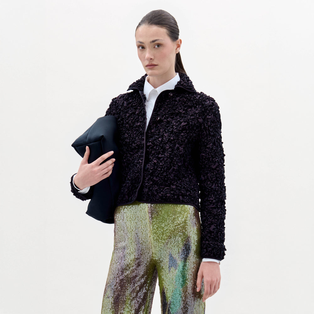 A woman stands against a plain background, wearing the textured black Jamari Jacket over a white shirt and shiny patterned pants, holding a folded black item in her left arm.