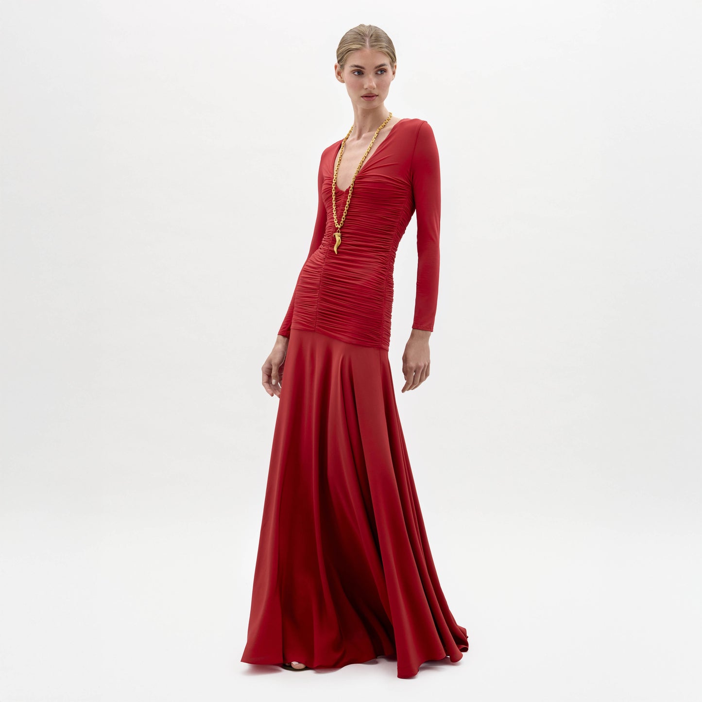 A woman models the Lina Dress, a long-sleeved, floor-length red gown with a deep V-neckline, paired with a gold necklace, against a plain white background.