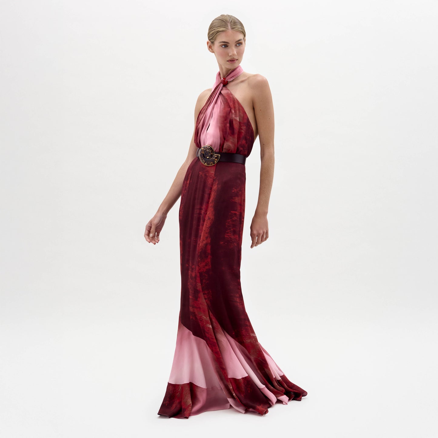 A woman stands against a plain white background wearing the sleeveless, floor-length Raisa Dress in red and pink with a wide black belt featuring a large floral buckle.