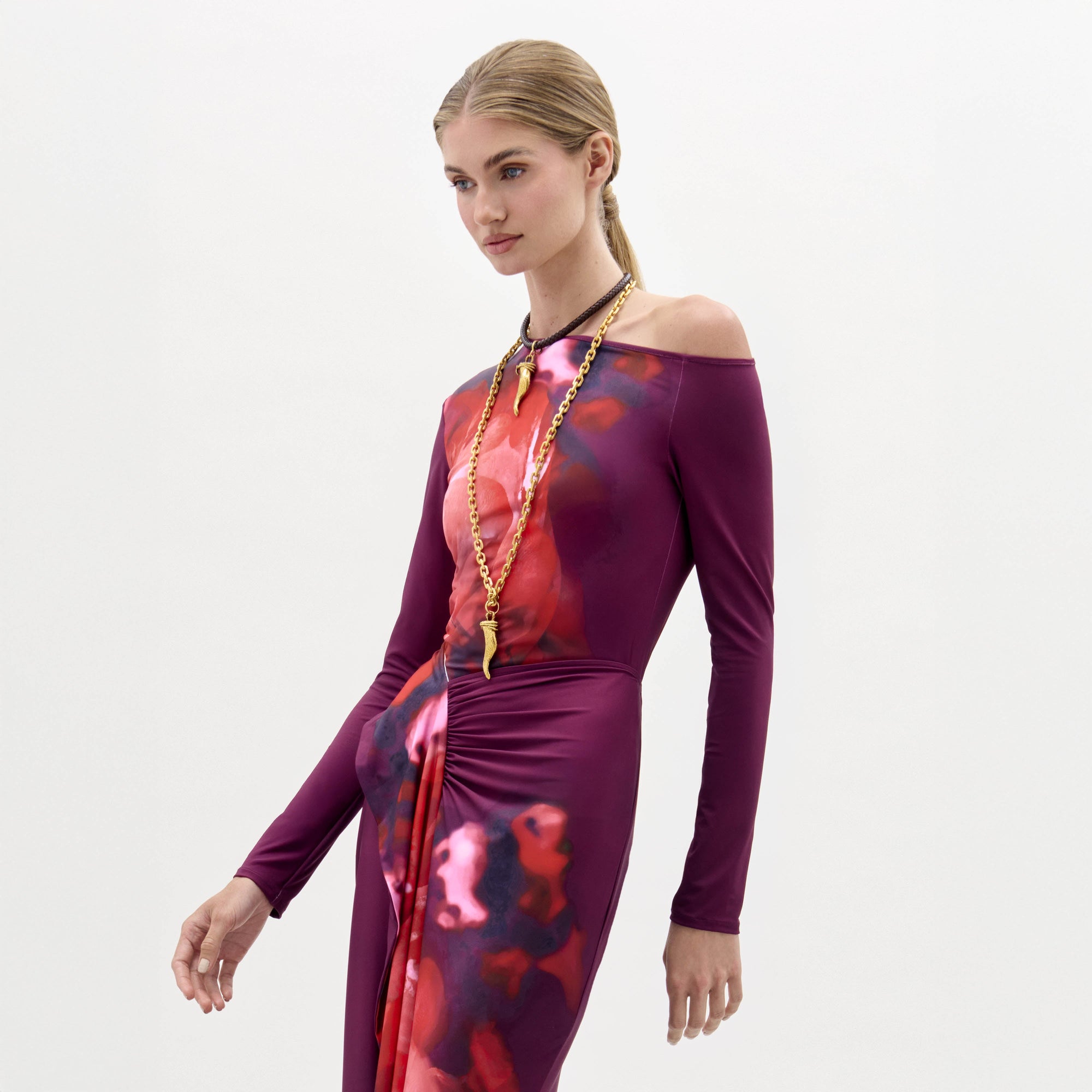 A woman wears the Sabra Blouse, a long-sleeve purple and red patterned top with an asymmetric neckline and a long gold necklace, standing against a plain white background.