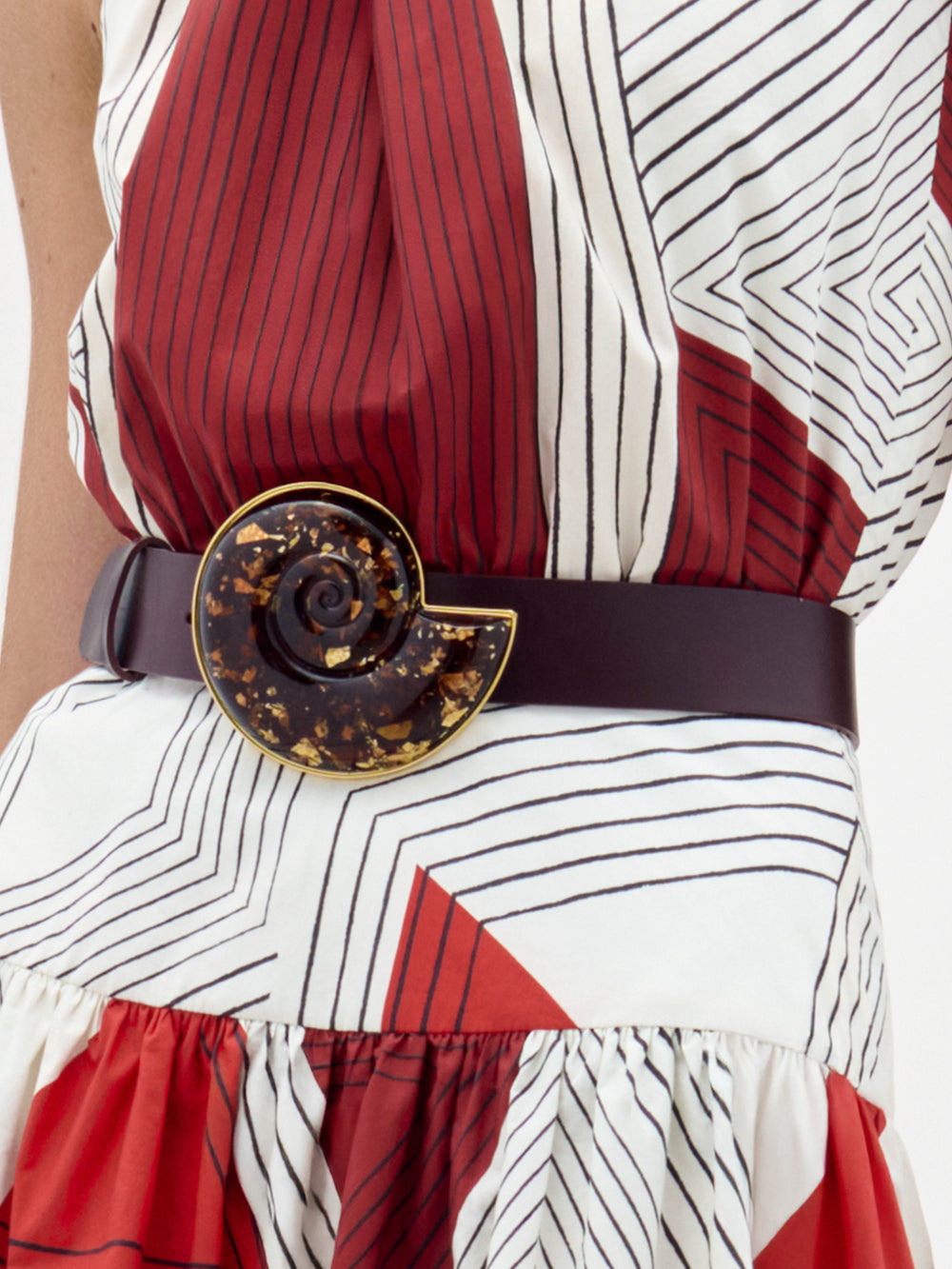 The Nautilus Belt is a dark maroon accessory with a large, glossy, spiral-shaped buckle adorned with amber and gold flecks.