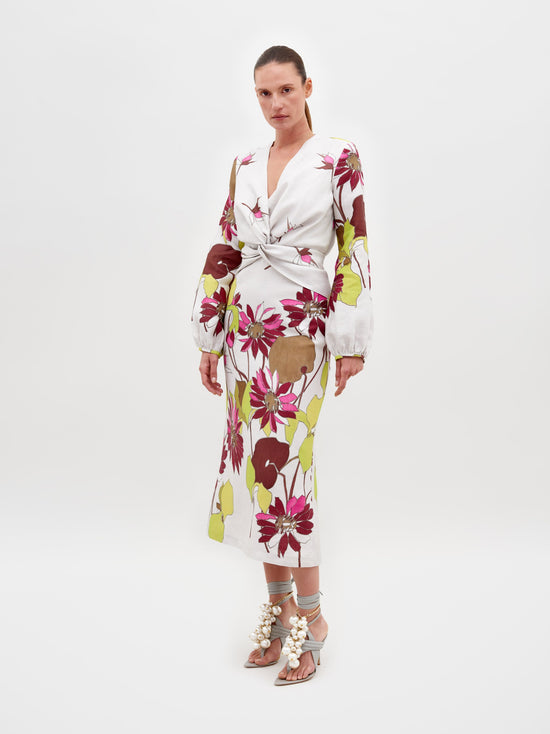 A woman stands against a plain background wearing the Battia Dress in Botanical Multifloral, which features a bold floral print, a cinched waist, and long sleeves. She is also wearing gray high-heeled sandals adorned with large pearl-like embellishments. The dress is available for pre-order now, with an estimated shipping date of February 15th, 2025.