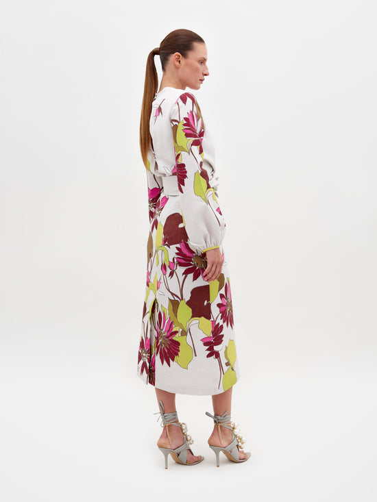 A woman stands against a plain background wearing the Battia Dress in Botanical Multifloral, which features a bold floral print, a cinched waist, and long sleeves. She is also wearing gray high-heeled sandals adorned with large pearl-like embellishments. The dress is available for pre-order now, with an estimated shipping date of February 15th, 2025.