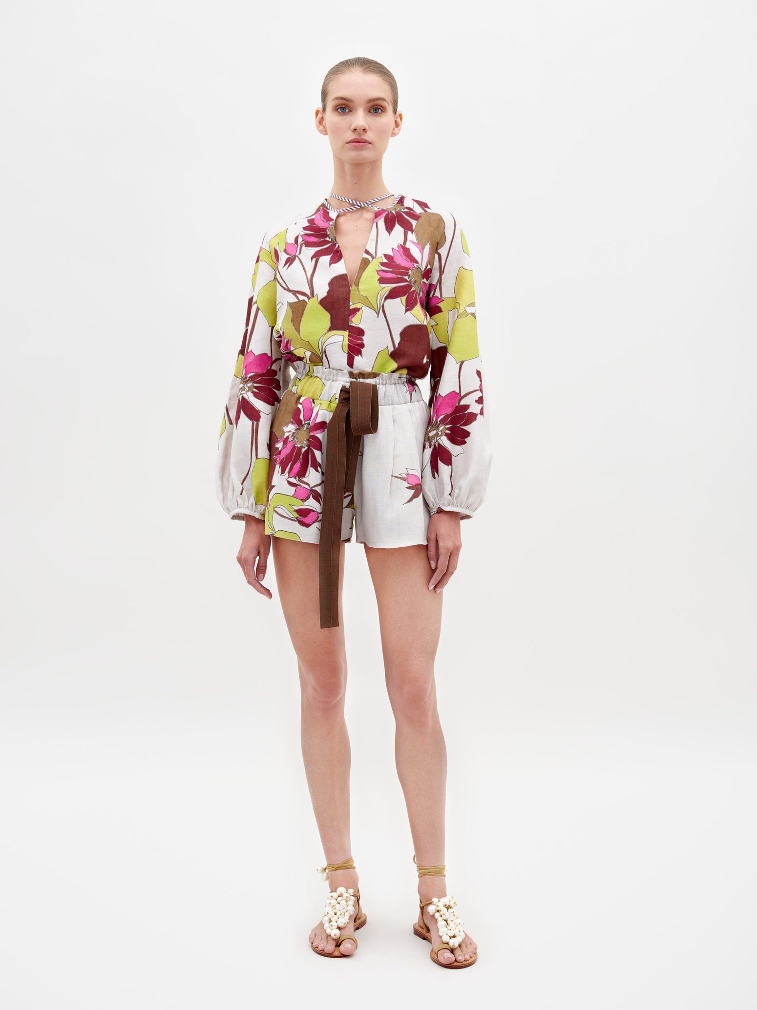 A person stands against a white background wearing the Giorgio Short Botanical Multifloral blouse, white shorts with a brown belt, and sandals. Pre-order this stylish ensemble now to ensure it arrives in time for February 15th, 2025.
