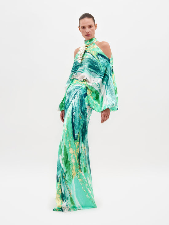 A person wearing the Claudia Skirt Green Marble dress, featuring a long-sleeved design with a shoulder cutout, poses against a plain white background. Pre-order now to ship by February 15th, 2025.